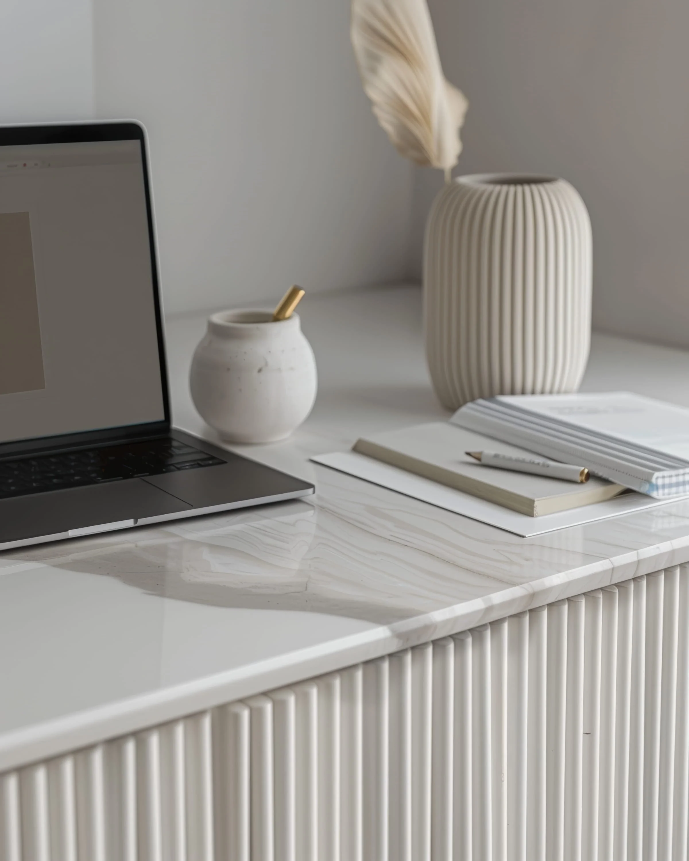 A white marble desk with a laptop, a small white vase, a large striped white vase with a dried feather, a notebook, a pen, and stacked papers.