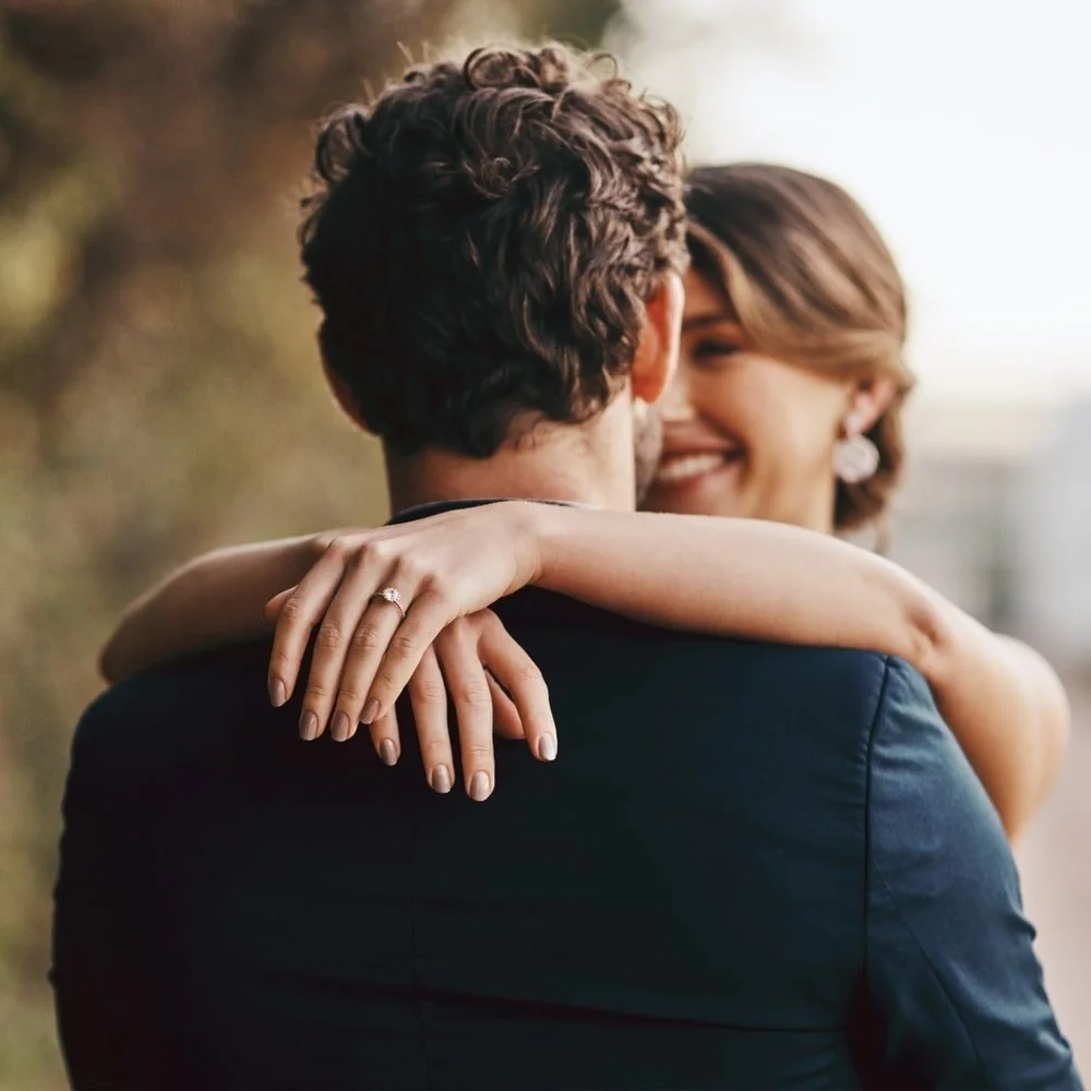 A couple embracing outdoors, with the woman smiling and showing an engagement ring on her finger.