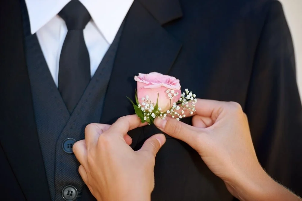 Person in a black suit and tie pinning a pink corsage with white baby's breath flowers onto another person wearing a black suit and white shirt.
