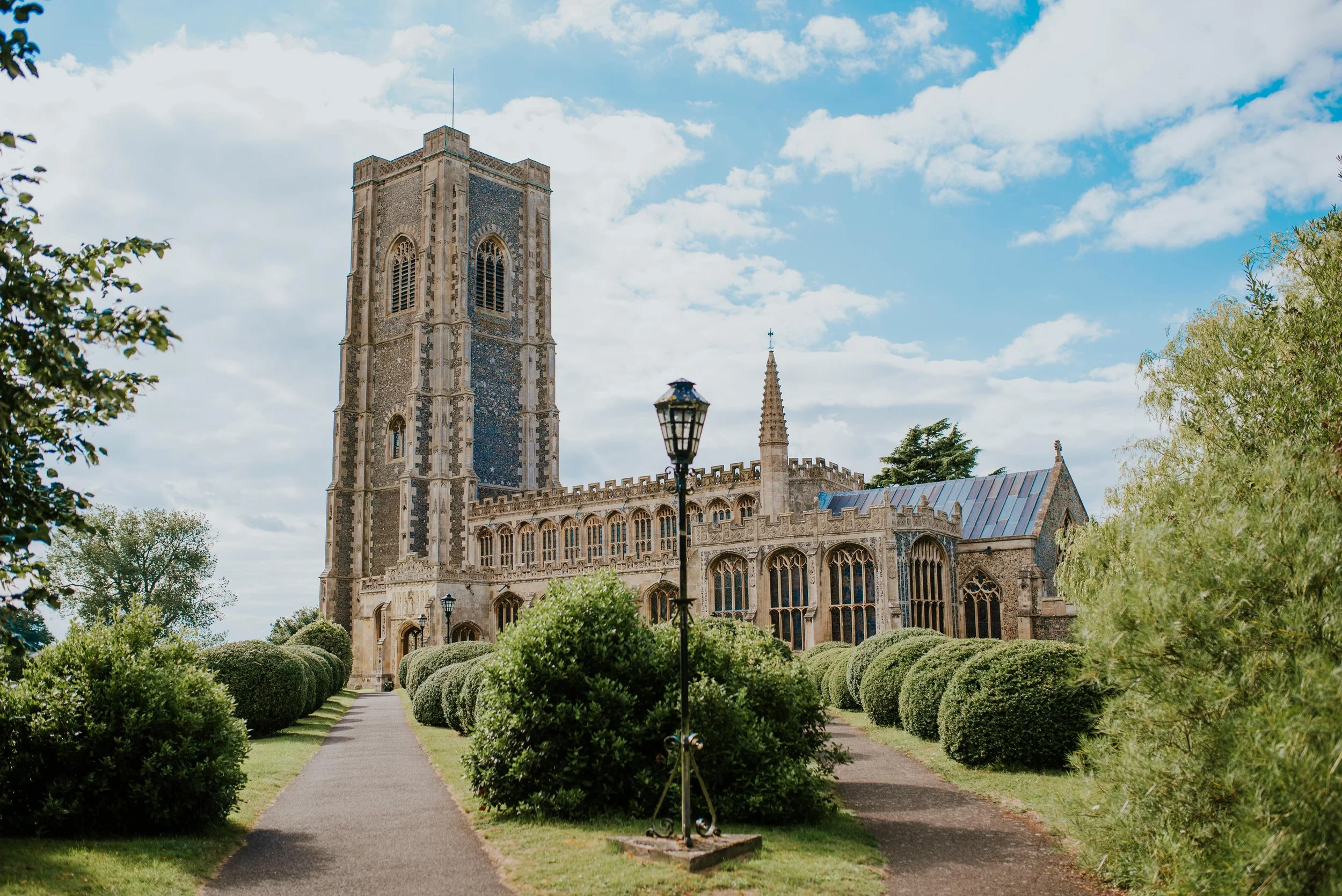 A historic stone church with a tall bell tower, arched windows, and decorative architectural details, surrounded by well-manicured bushes and trees under a partly cloudy sky.