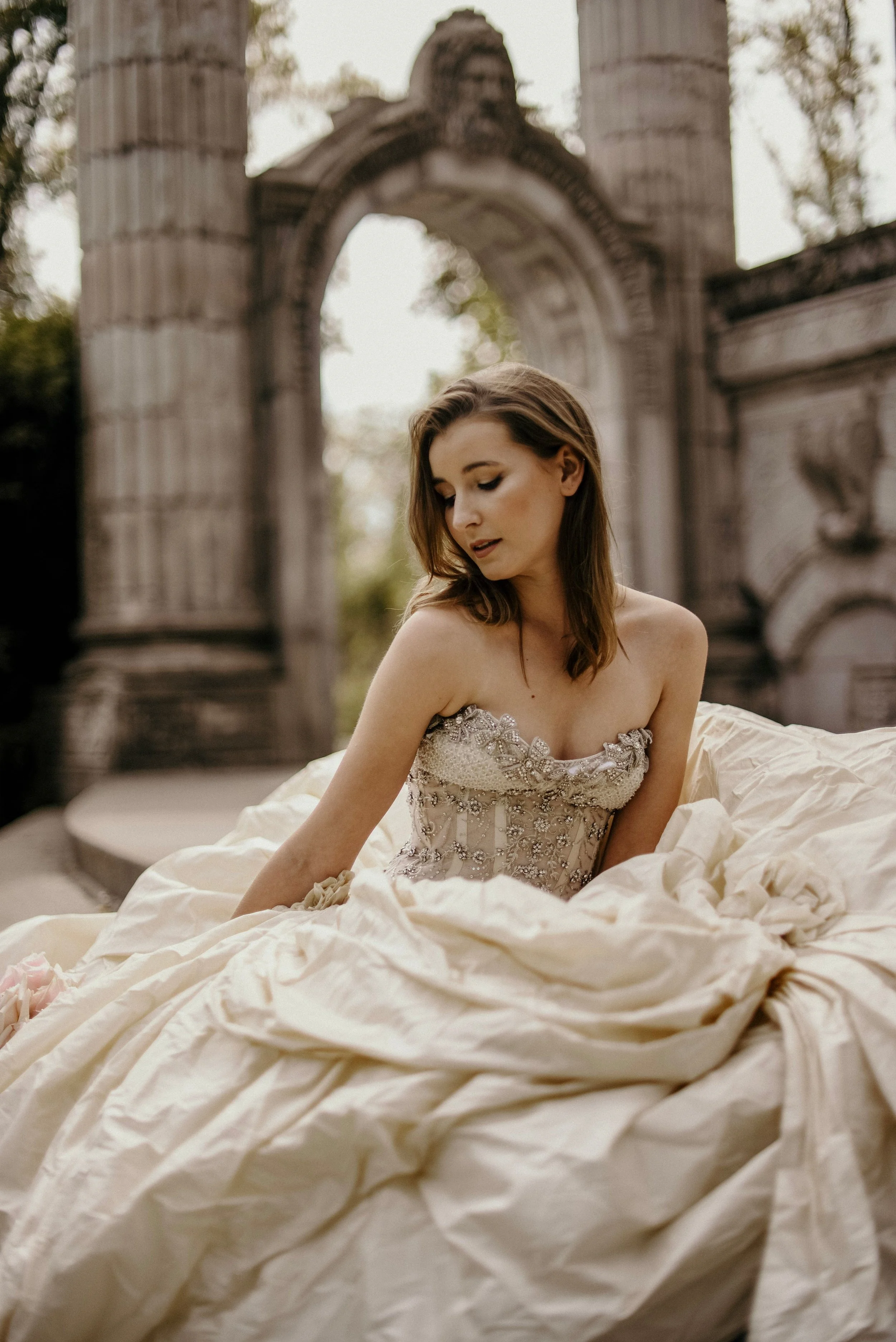 A young woman in an elaborate, strapless, vintage-style wedding dress with floral embroidery, sitting outdoors in front of a stone archway with classical columns and sculptures, surrounded by trees.
