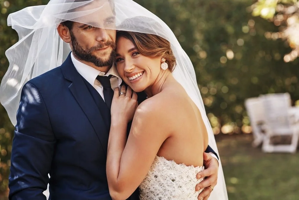 Happy bride and groom posing outdoors, with the bride smiling and leaning into the groom, wearing a wedding dress and veil, and the groom in a navy blue suit with a white shirt and black tie.