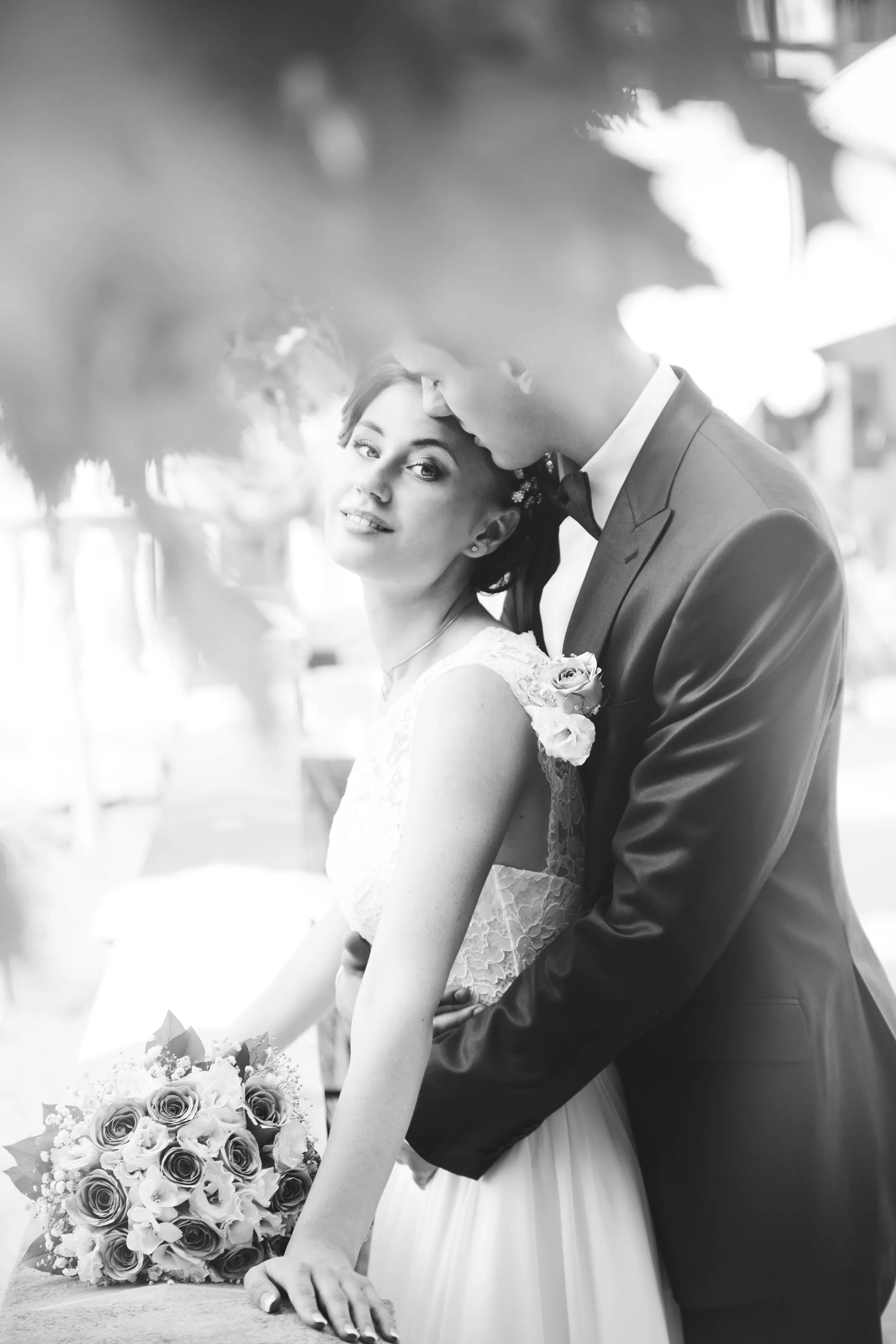 Black and white photo of a bride and groom on their wedding day, with the groom gently kissing the bride's forehead while she holds a bouquet of roses.
