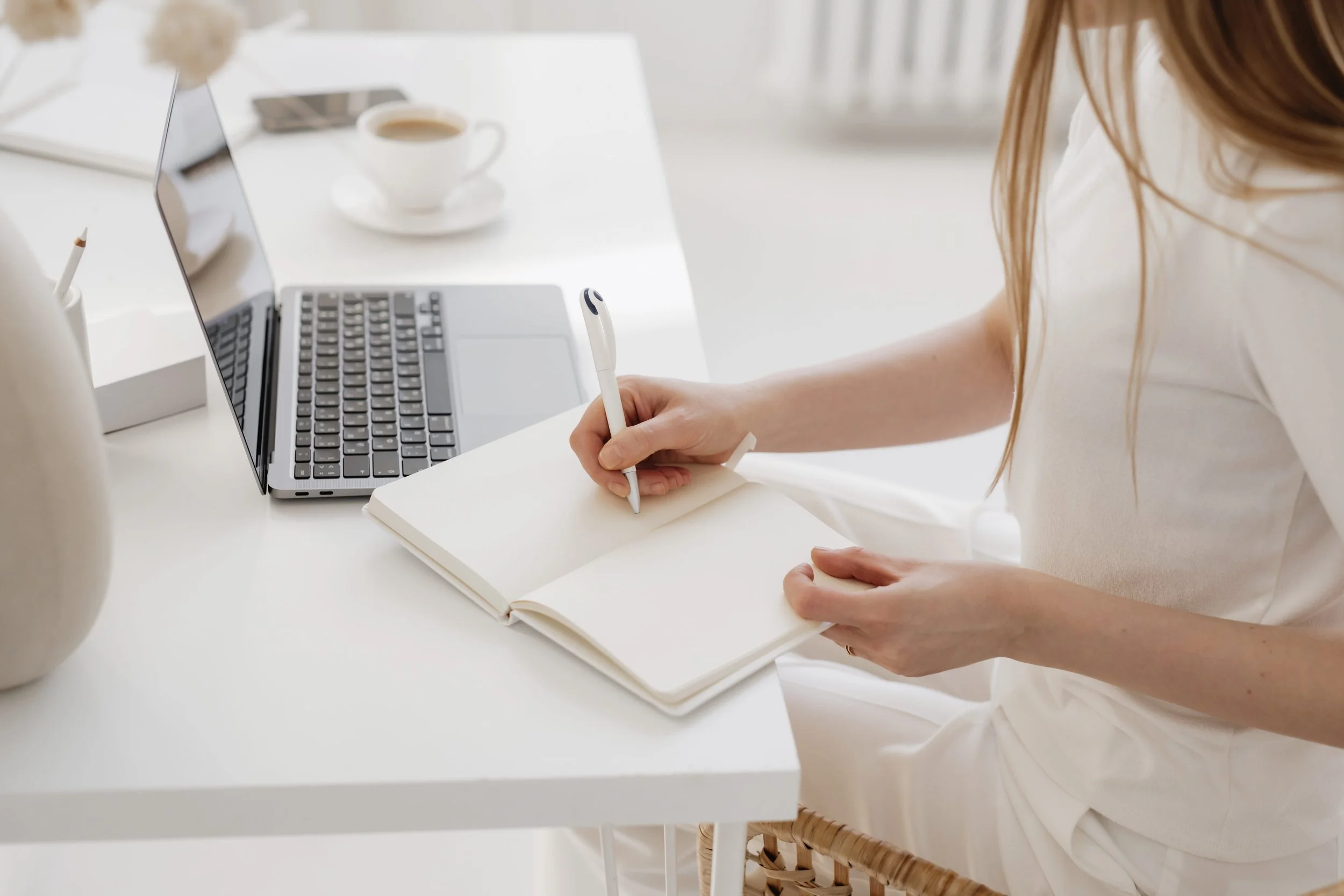 A person sitting at a white table, writing in a notebook with a pen, an open laptop, a cup of coffee, and a smartphone on the table.