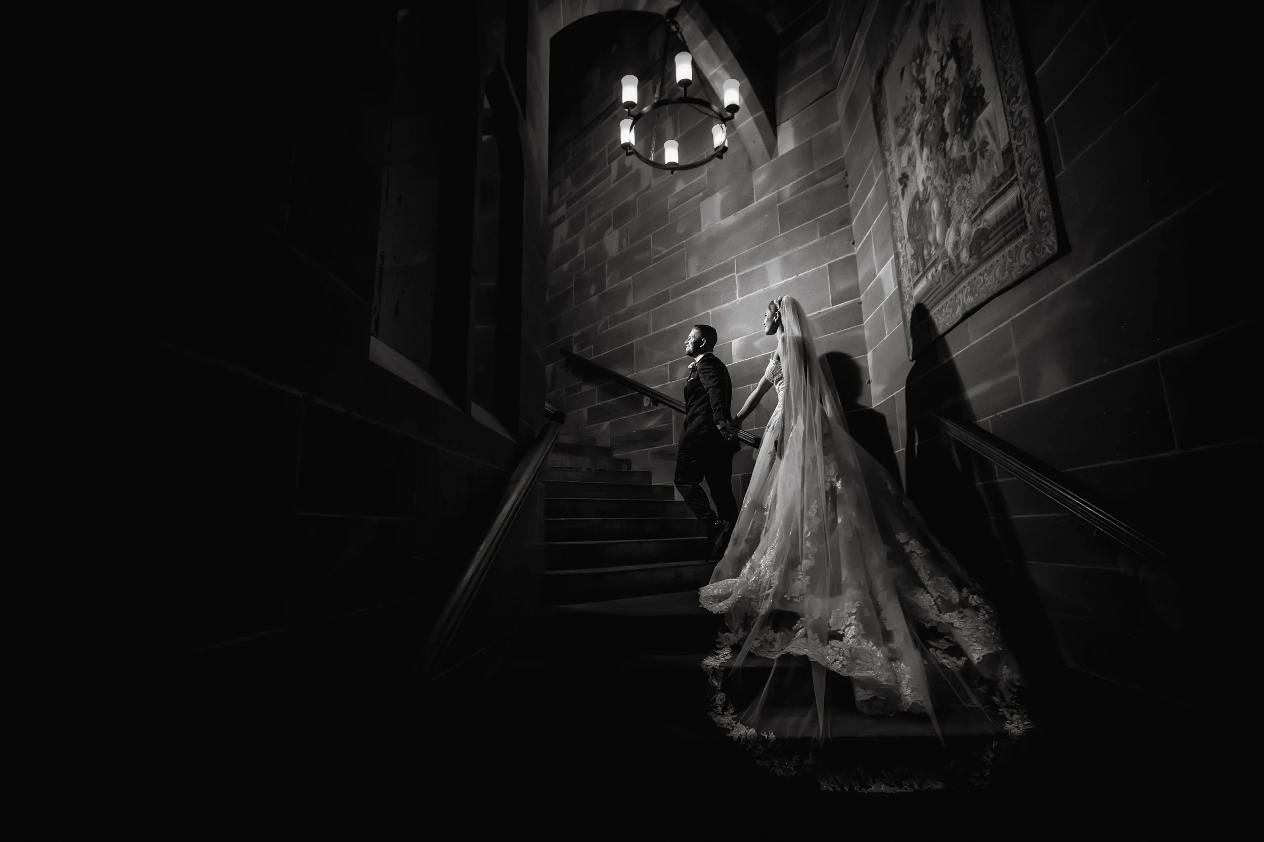 A bride and groom holding hands while ascending stairs in a dark, Gothic-style interior with a large chandelier and medieval tapestry, captured in black and white.