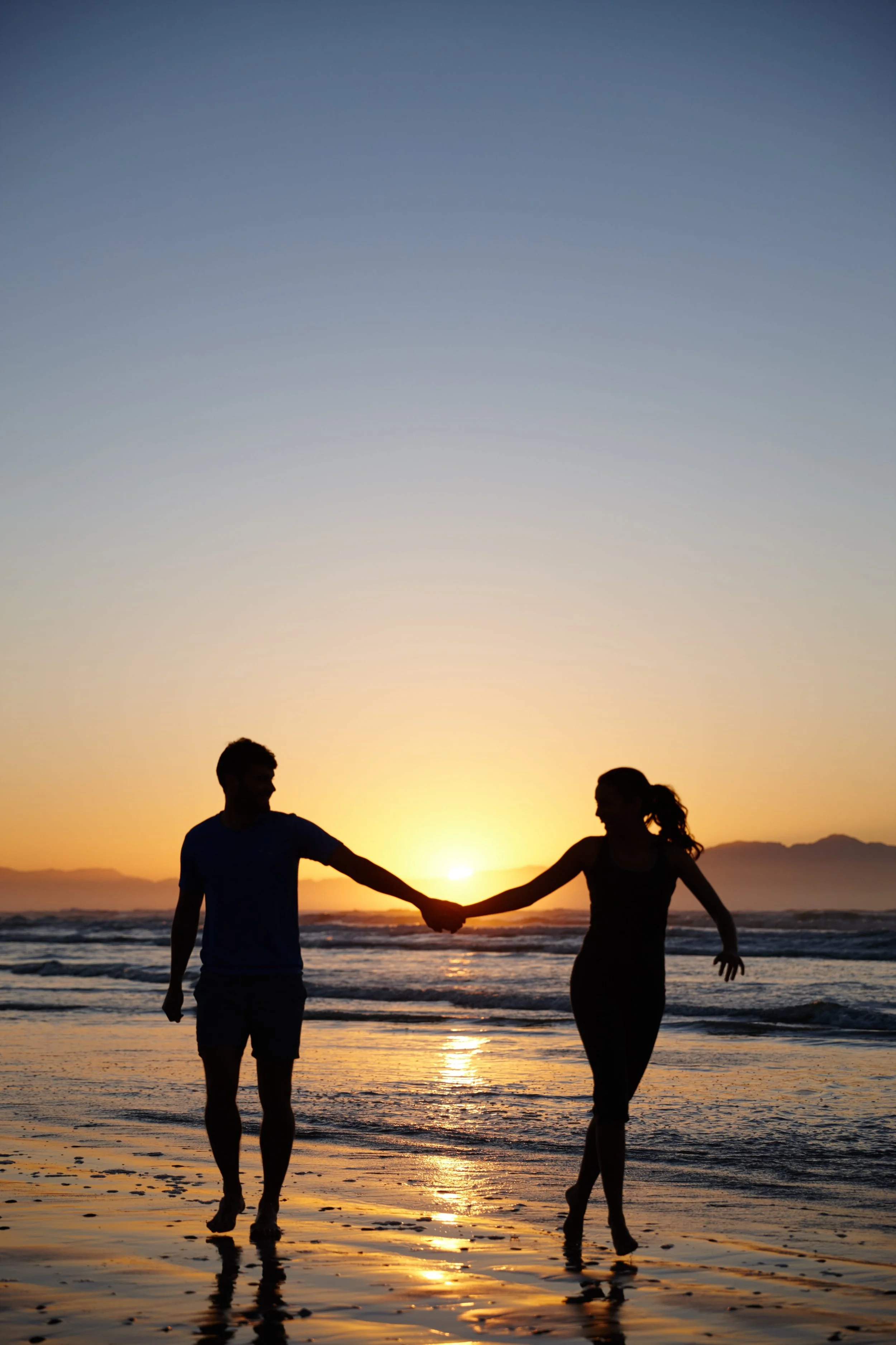 A silhouette of a man and woman holding hands and walking along the beach during sunset.