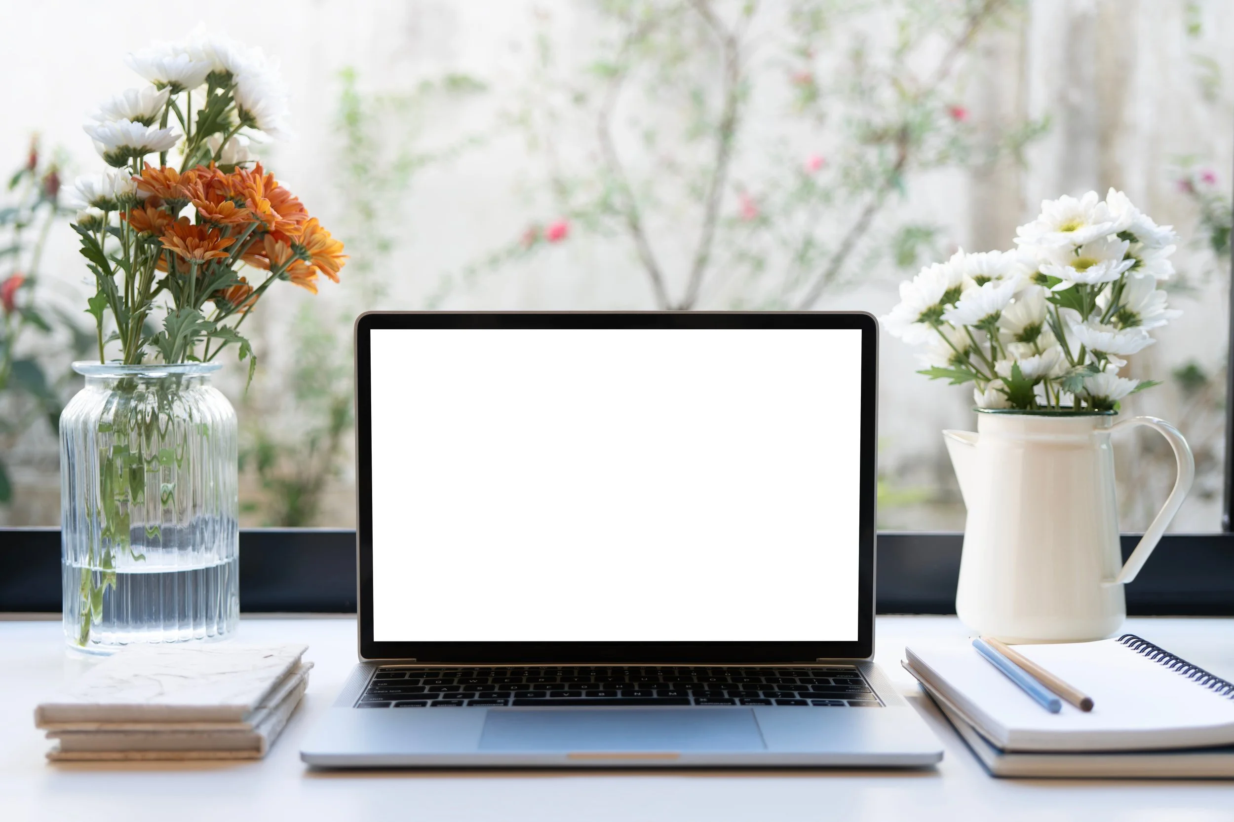 Laptop on a white desk with a glass vase of orange and white flowers on the left and a white pitcher of white flowers on the right, with notebooks and pens in front.