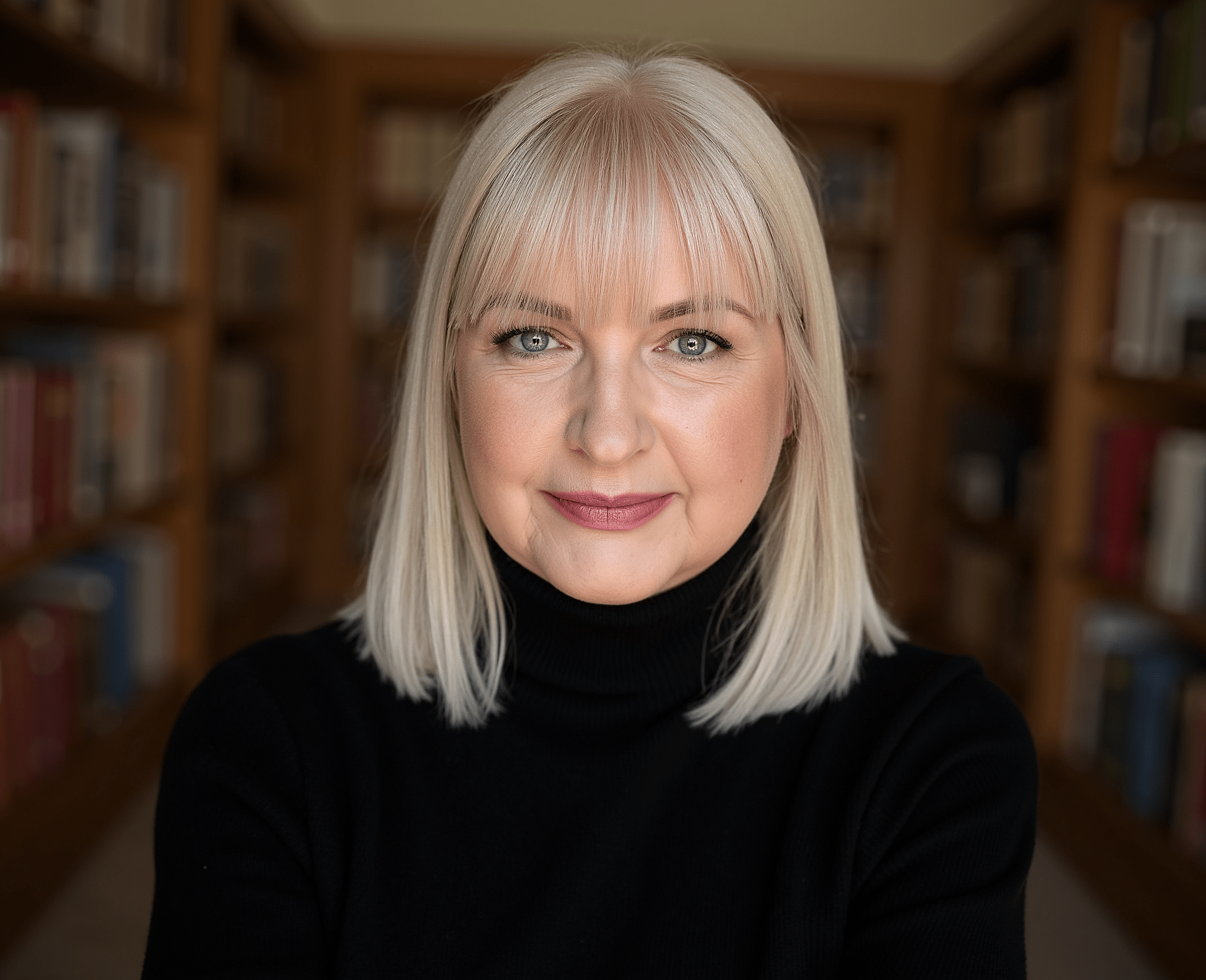 A smiling woman with platinum blonde hair and piercing blue eyes, wearing a black turtleneck, standing in front of a blurred bookshelf.