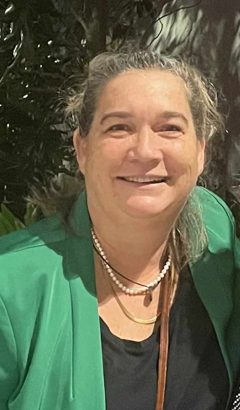 Close-up of a smiling woman with gray hair styled back, wearing a green blazer, black top, pearl necklace, and gold jewelry, standing in front of a dark, leafy background.