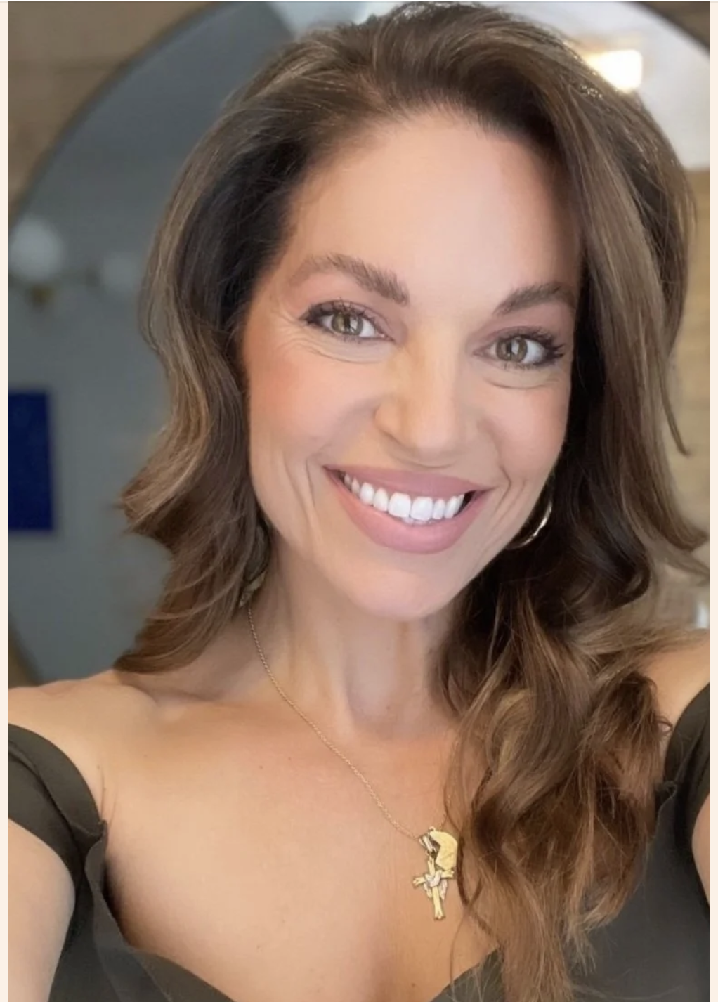 Close-up of a smiling woman with long wavy brown hair, wearing a black top, gold cross necklace, and hoop earrings.