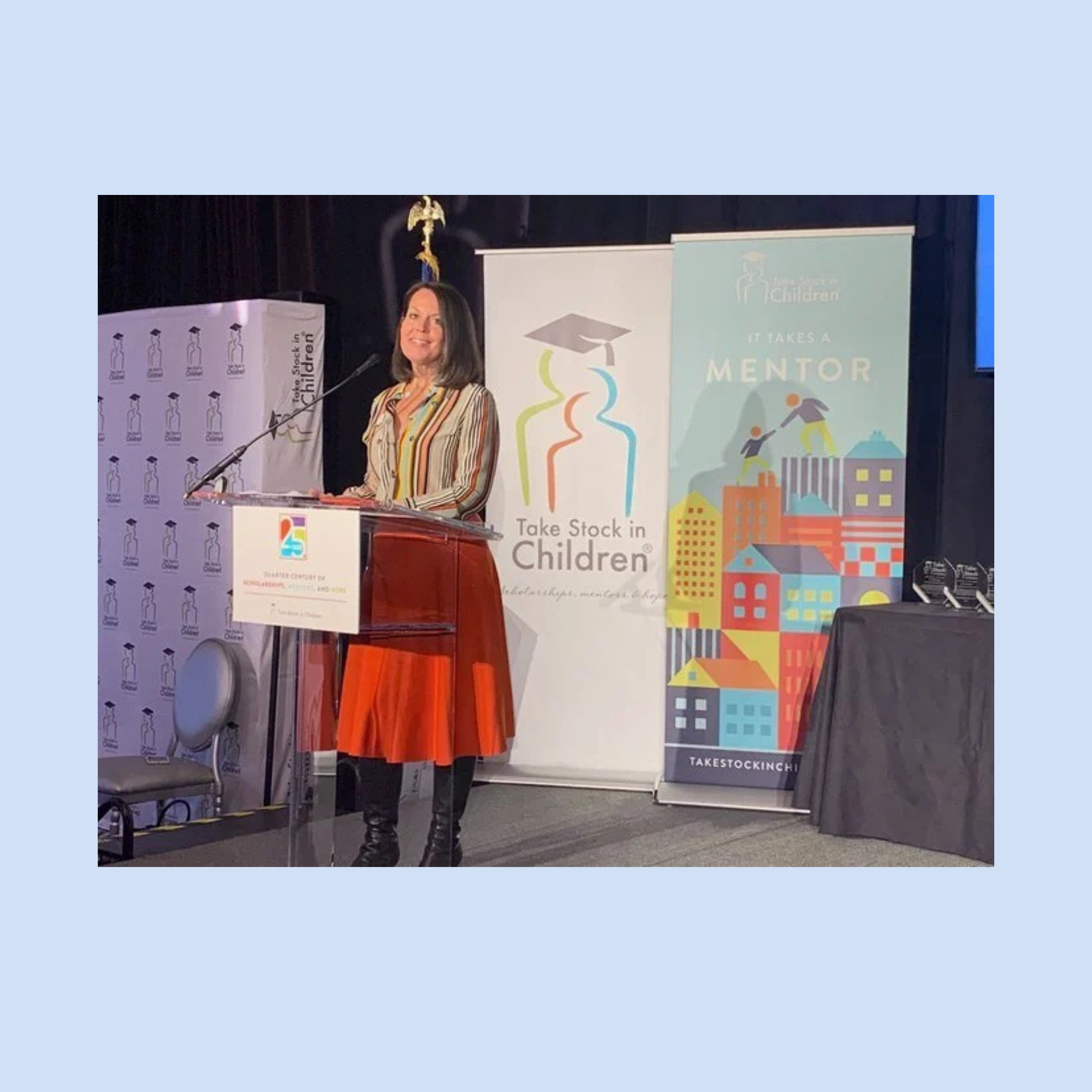 Woman speaking at a podium during an event, with banners and signage in the background.