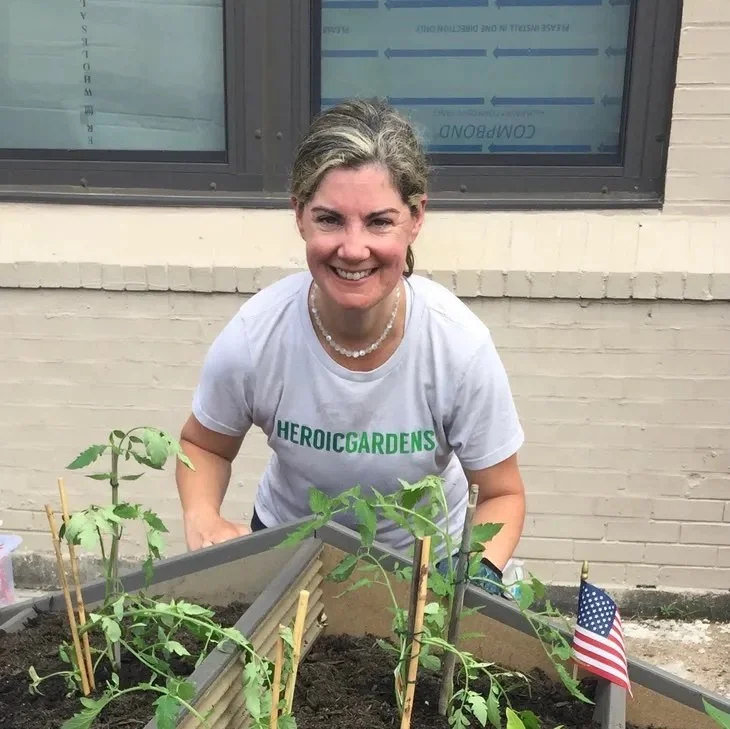 Woman tending to a raised garden bed with young plants, smiling outdoors.