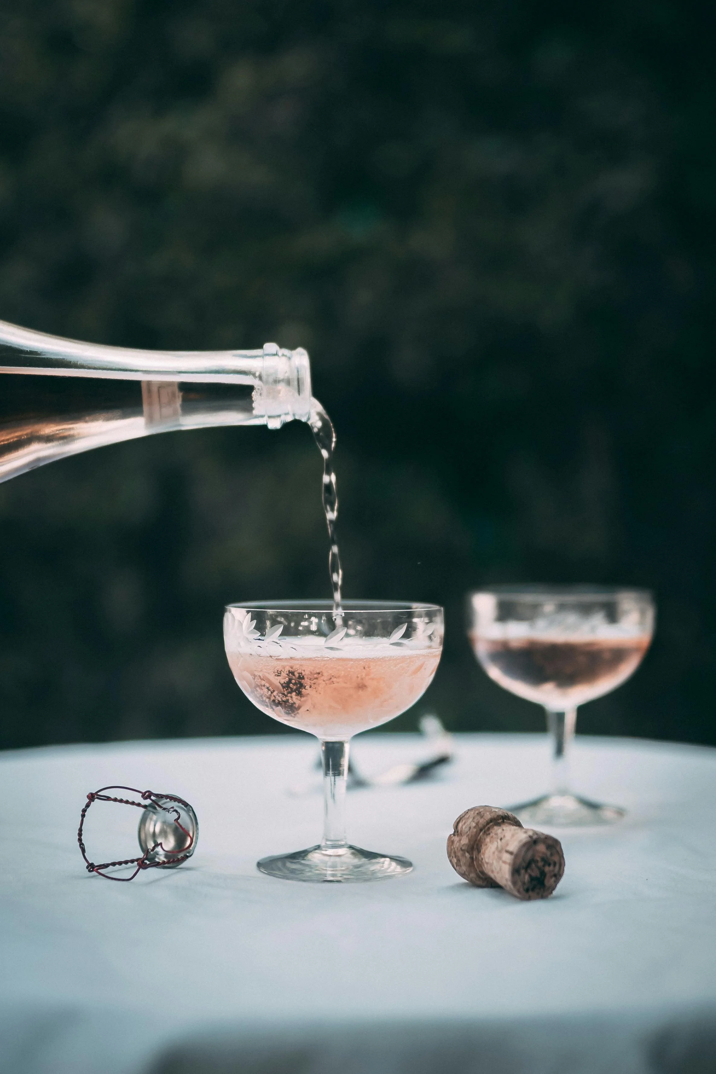 Zwei cocktailartige Getränke in eleganten Gläsern, die auf einem hellen Tuch auf einem Tisch stehen, dazu eine Korken und eine kleine Handlupe, der Hintergrund ist unscharf und dunkel.