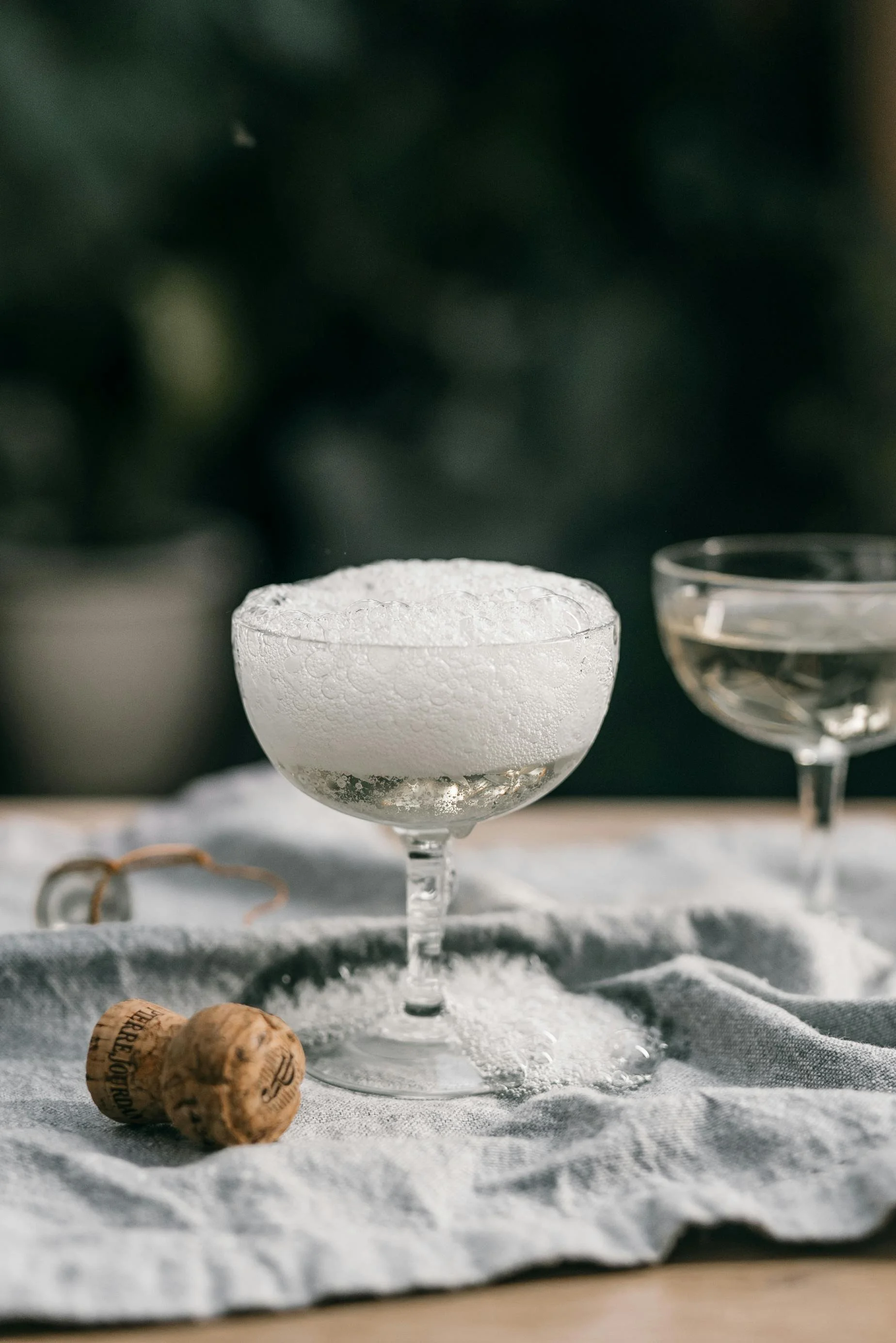 Zwei Champagnergläser mit Schaum, eins ist gefüllt, das andere ist teilweise sichtbar, auf einem grauen Tuch. Zwei Korken liegen daneben. Unschärfer Hintergrund.