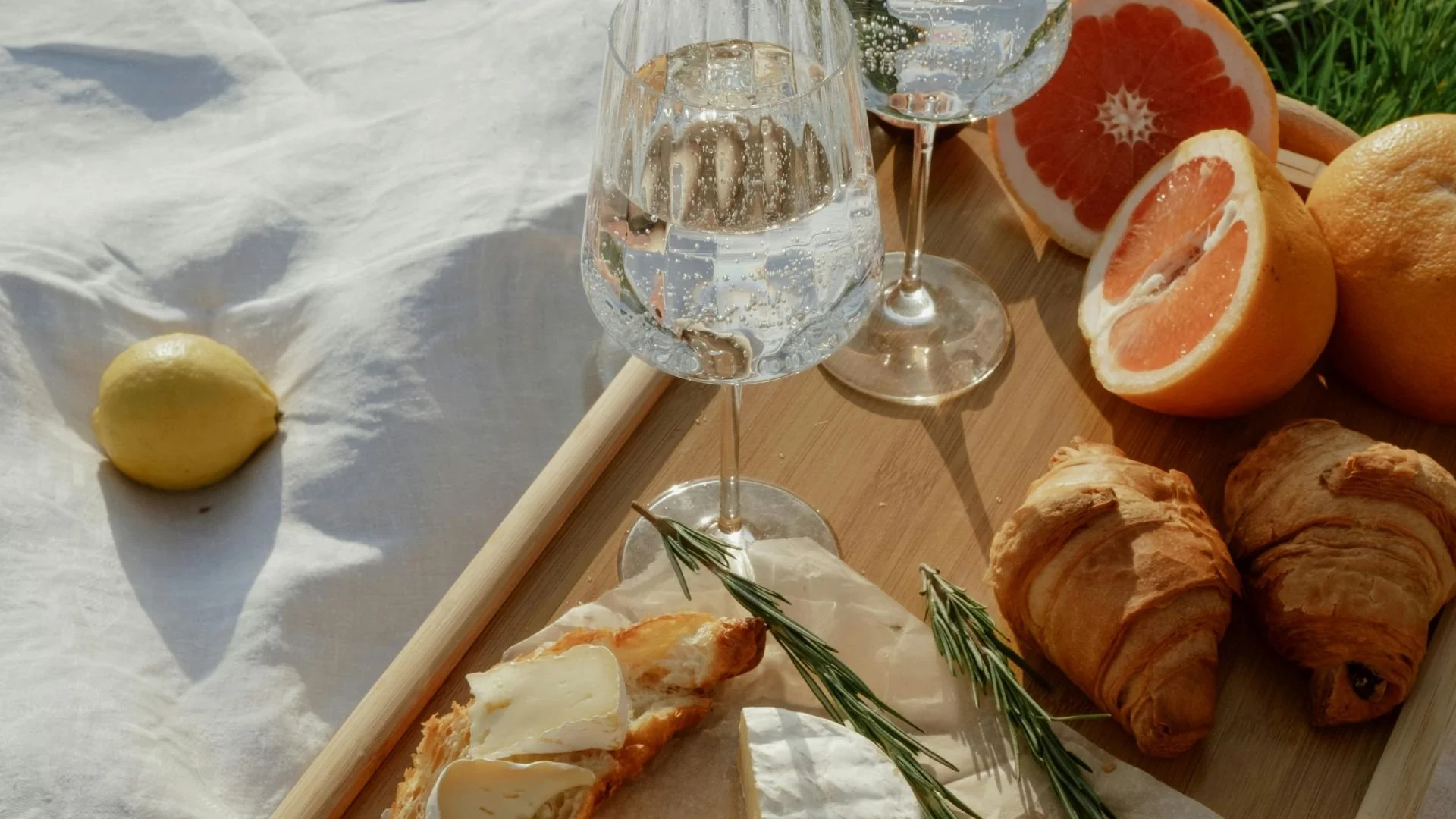 Ein Picknicktisch mit zwei Gläsern Wasser, einer halben Grapefruit, Zitronen, Croissants, einem Stück Käse, Rosmarin, einem Stück Pizza mit Käse, und einer anderen Frucht auf grünem Gras