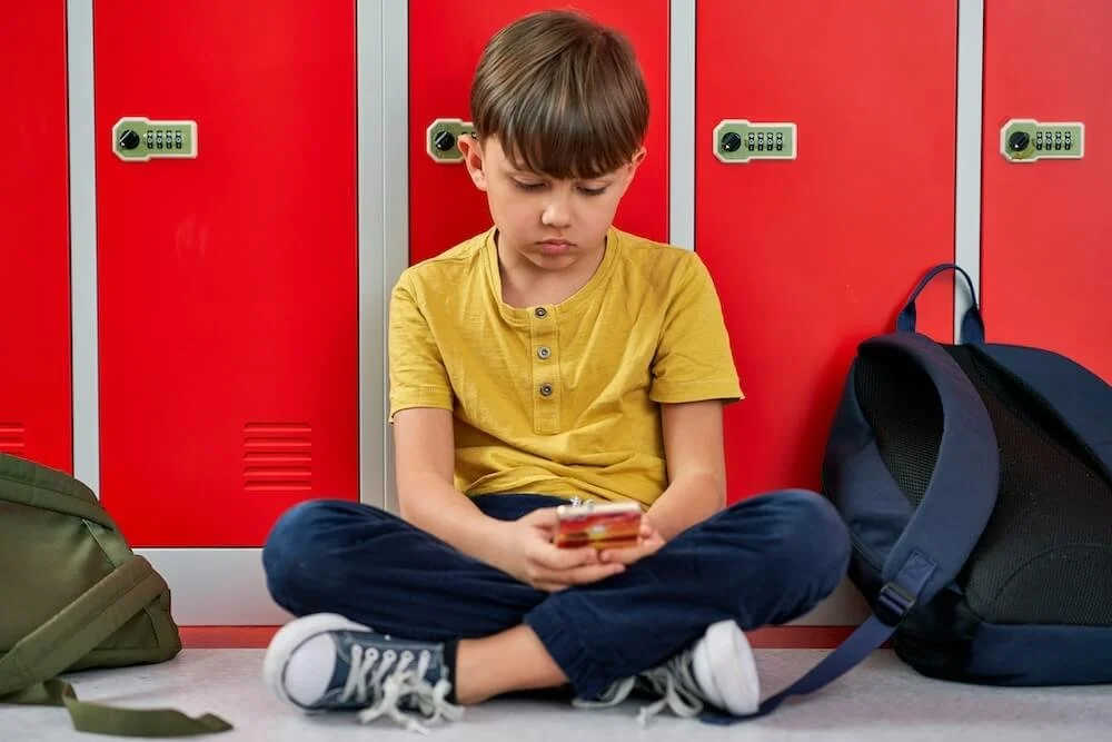 Sad boy sitting on the floor against lockers looking at a cell phone. Connect your child with a child therapist in New York, NY to help them cope with bullying and navigate the effects in healthy ways.