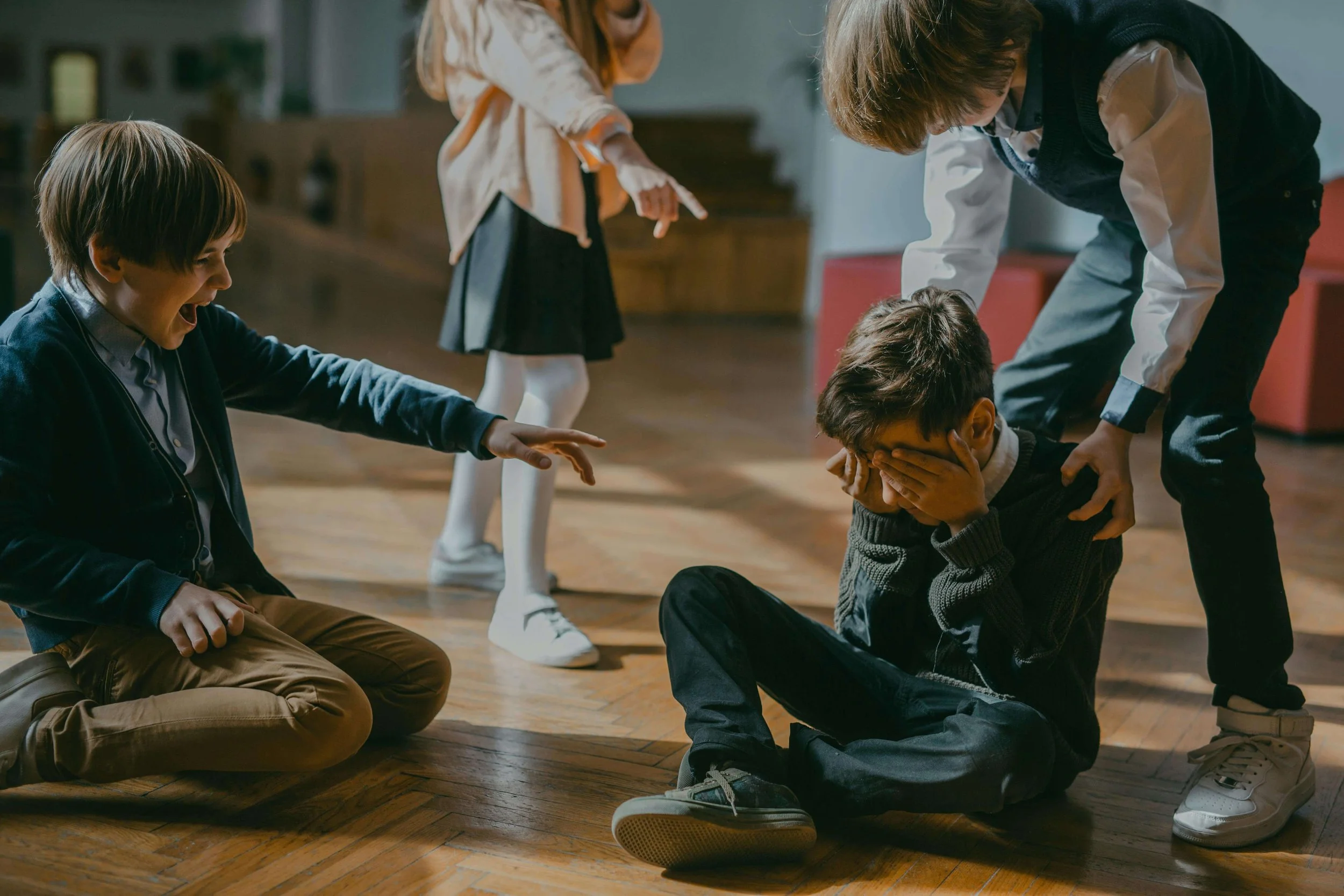 Sad boy sitting on the ground covering his eyes as other children point and laugh. Find support for your child and help them heal from school bullying with the compassionate support of a child therapist in New York, NY.