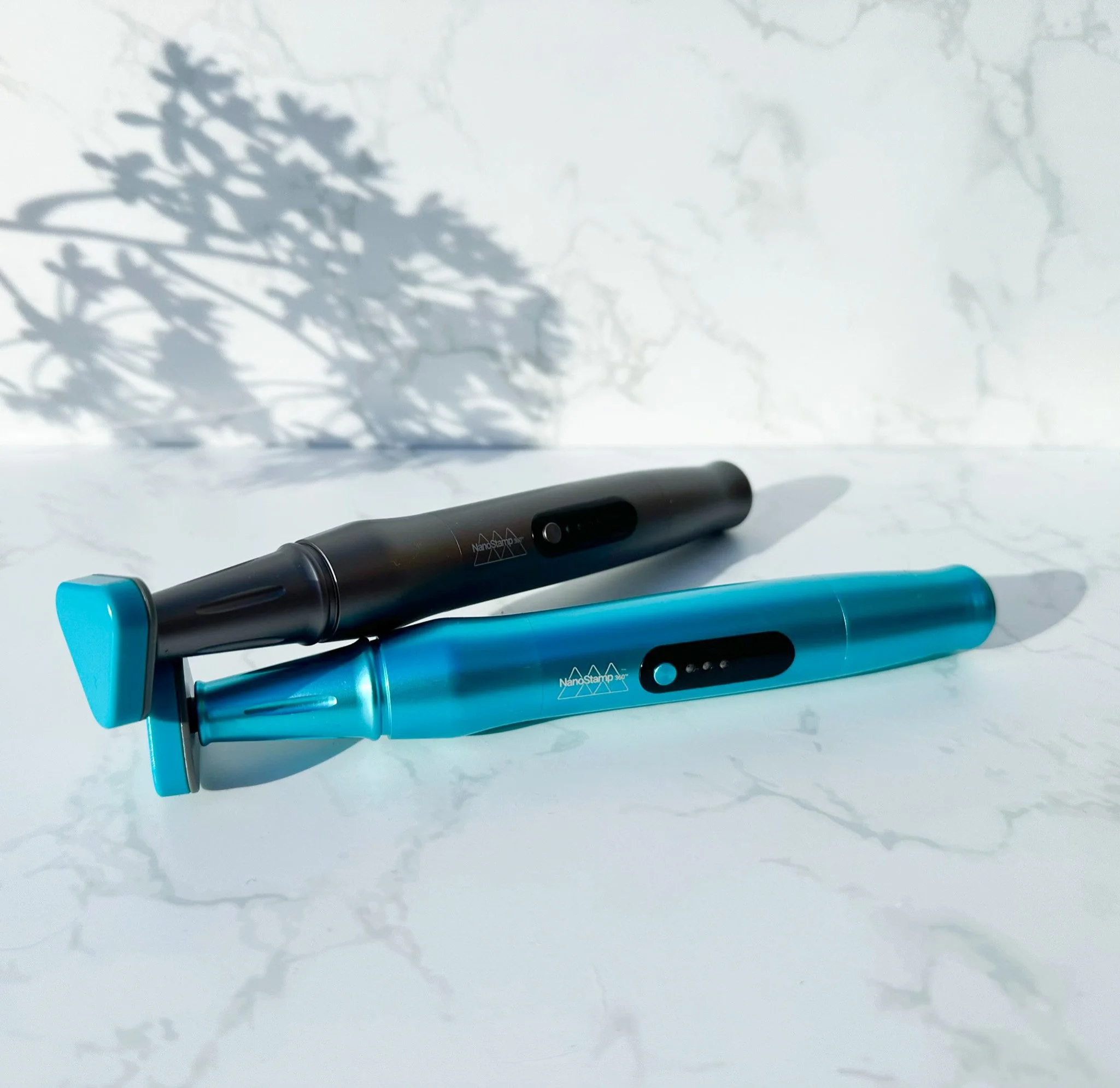 Two blue and black NanoStamp 360 devices placed on a white marble surface with shadows of leaves in the background.