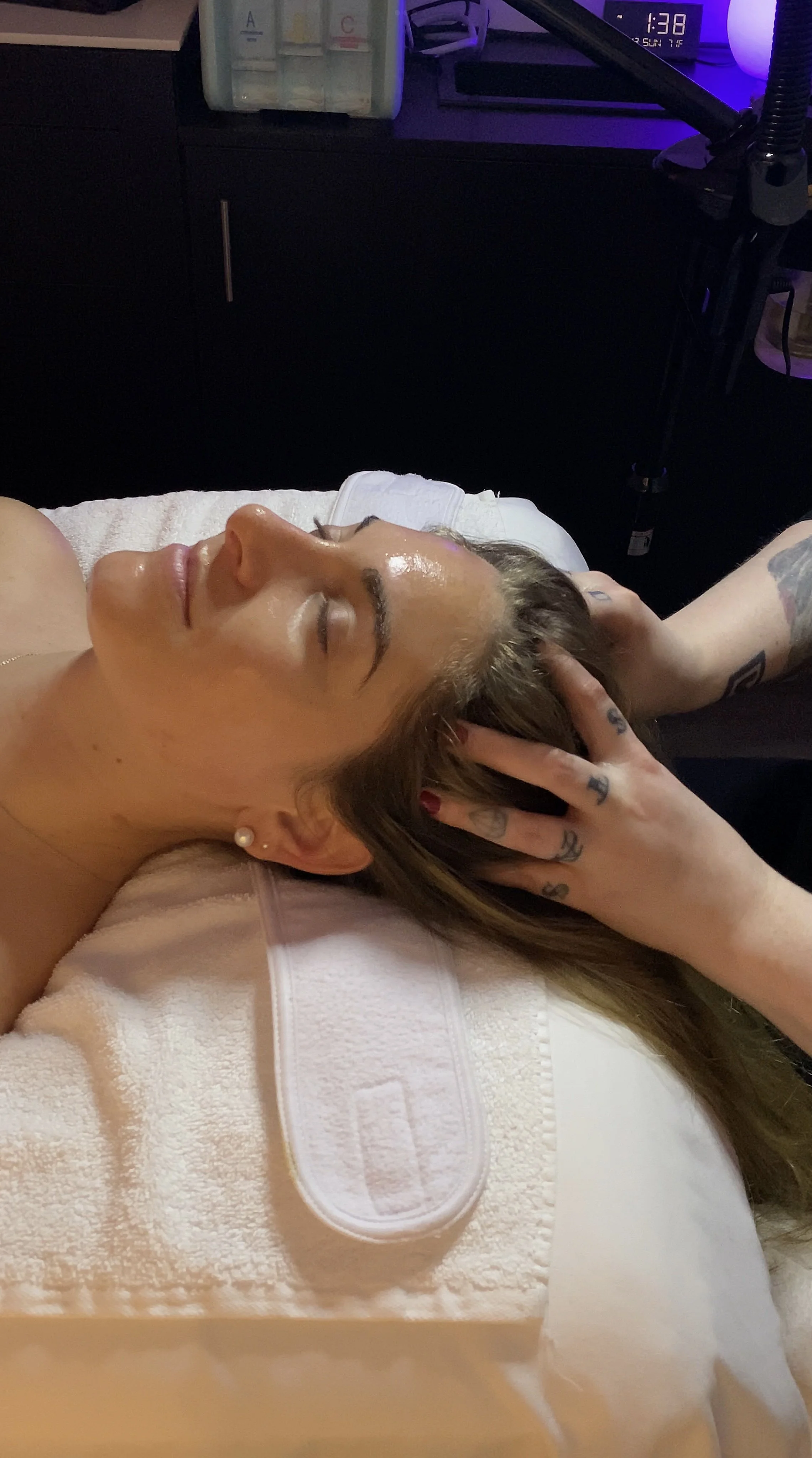 A woman with tattoos lying on her back with her eyes closed, receiving a massage or facial treatment, in a dimly lit room with purple lighting.