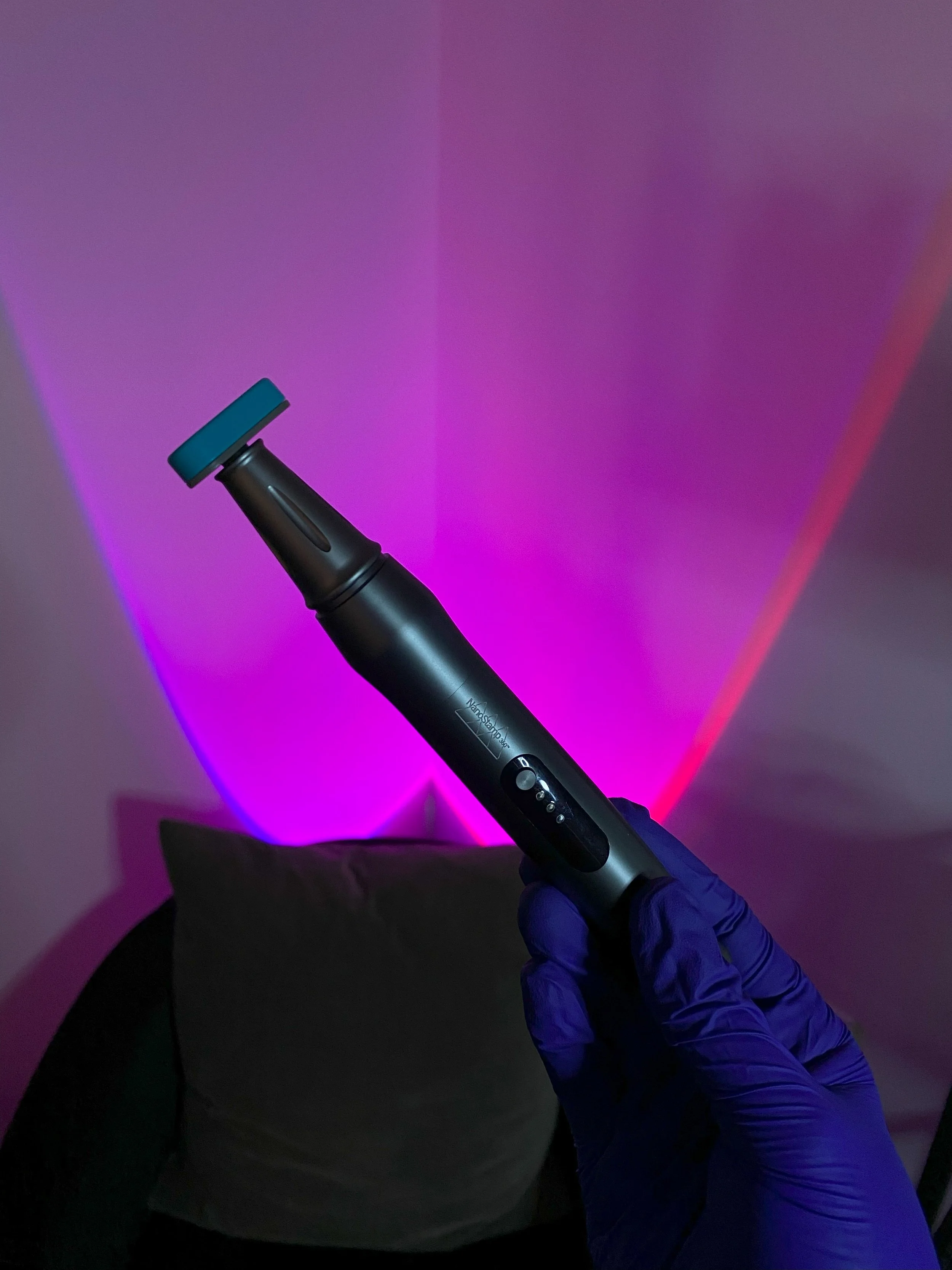 A person wearing a blue glove holding a black handheld device with a large roller head, set against a background with pink and purple lighting.