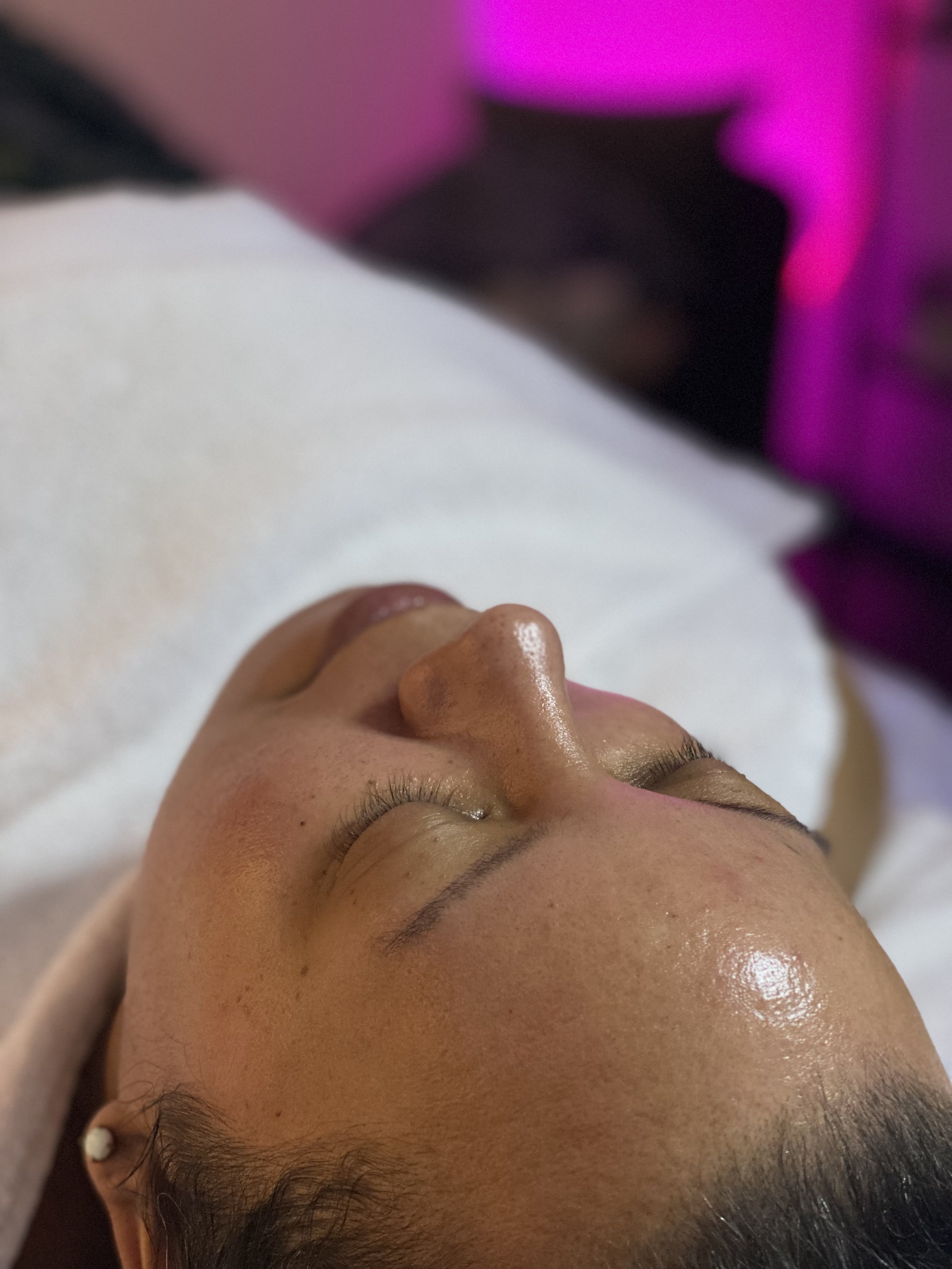 Close-up of a woman lying down with eyes closed, receiving a facial treatment, with a blurred pink and purple light in the background.