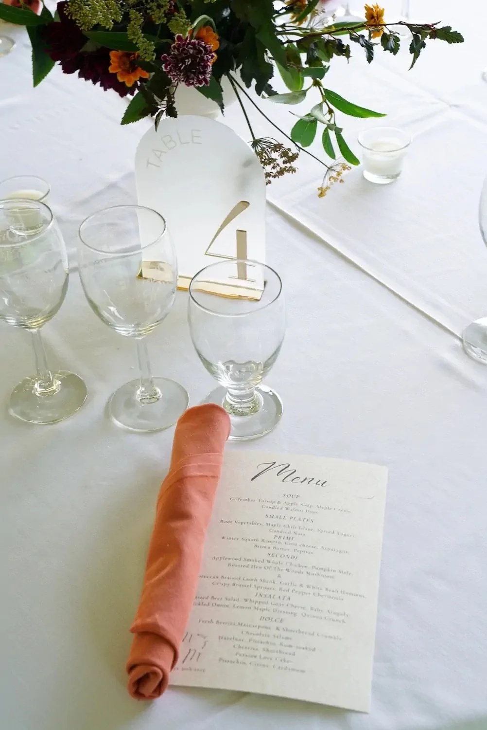 Elegant dining table setup with a floral centerpiece, clear wine glasses, a menu, and a white table number card reading '1' on a white tablecloth.