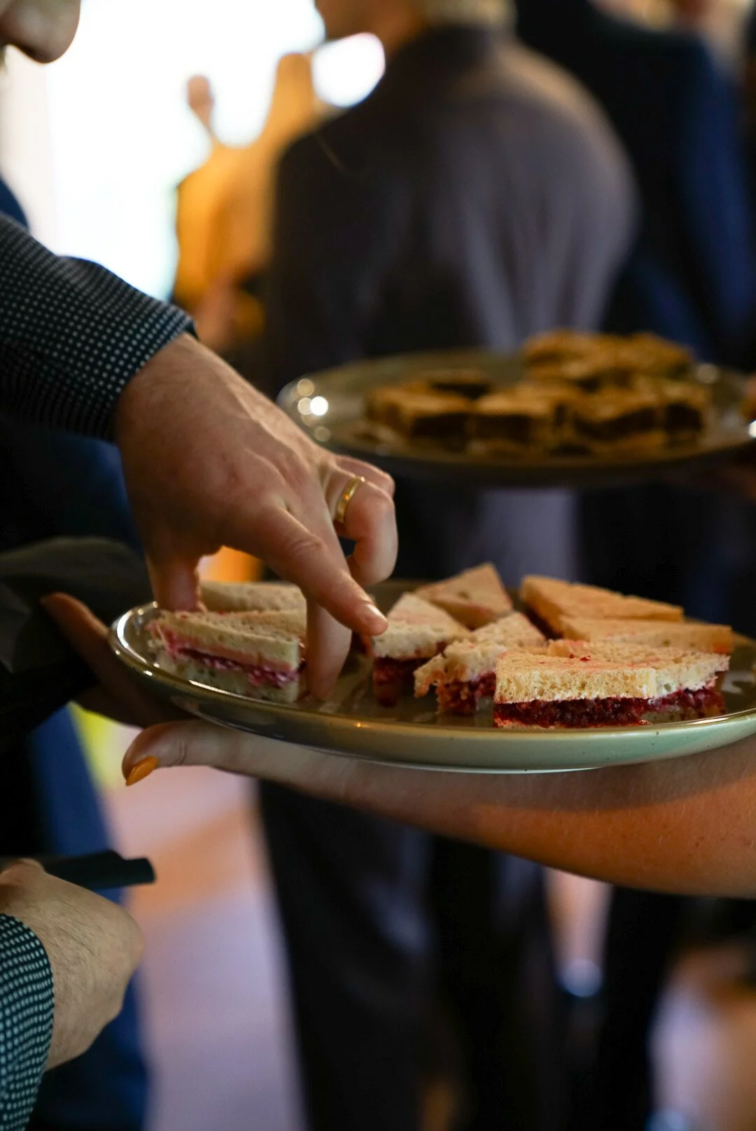 Person reaching for a plate of assorted sandwiches at a buffet, with other people in the background.