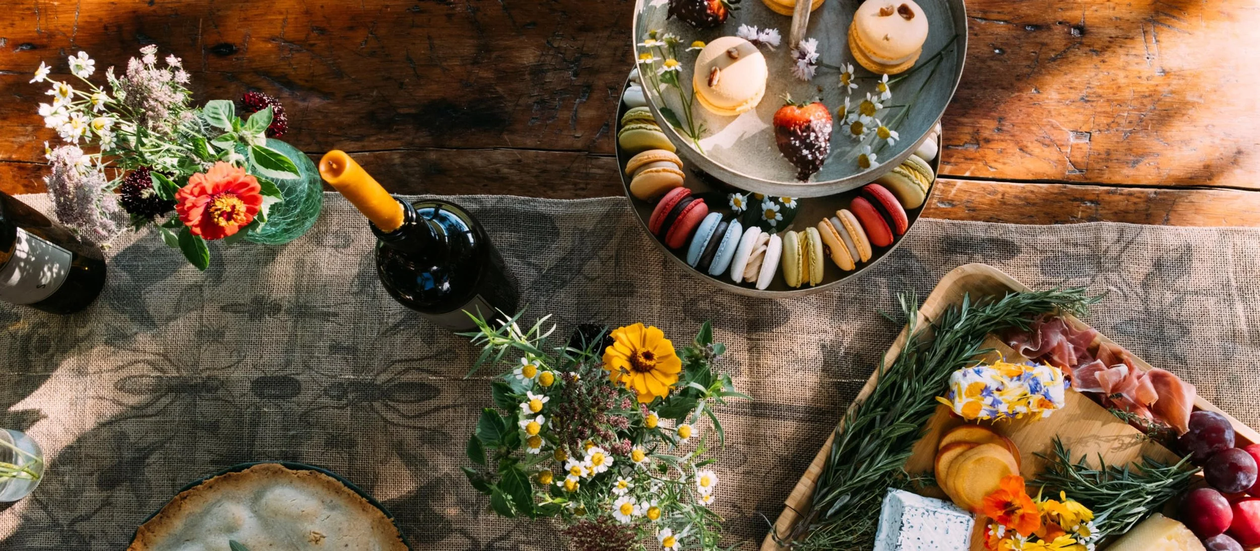 A table set for a celebration with flowers, wine, cheese, fruit, and colorful desserts, including macarons and strawberries.