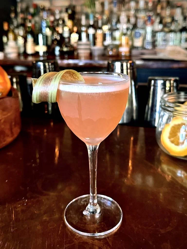 A pink cocktail in a coupe glass garnished with a lime peel, on a dark wooden bar counter with blurred bottles in the background.