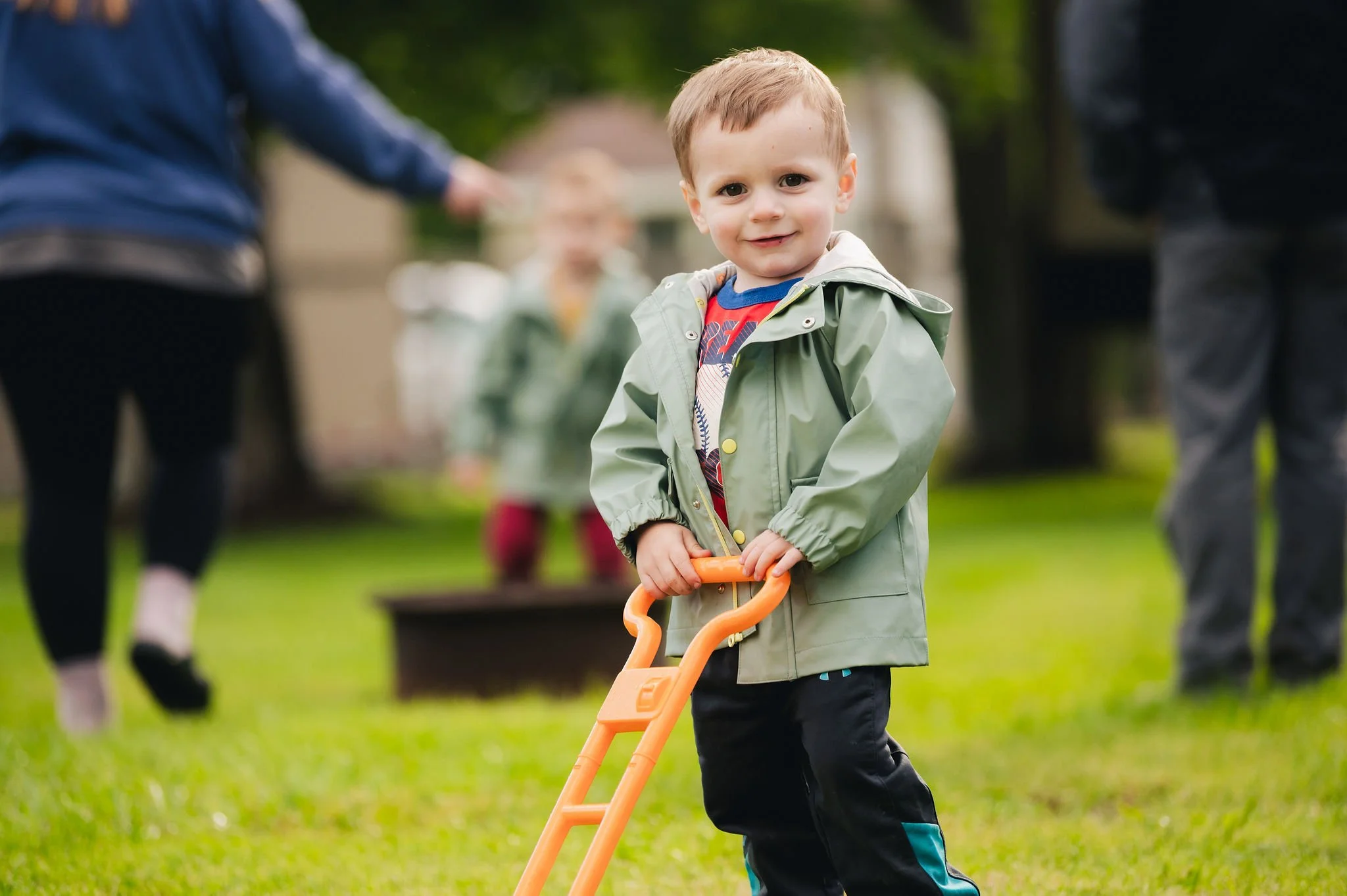 A young boy with brown hair wearing a green jacket and black pants holding an orange toy shovel in a grassy backyard, with blurred children and adults playing in the background.