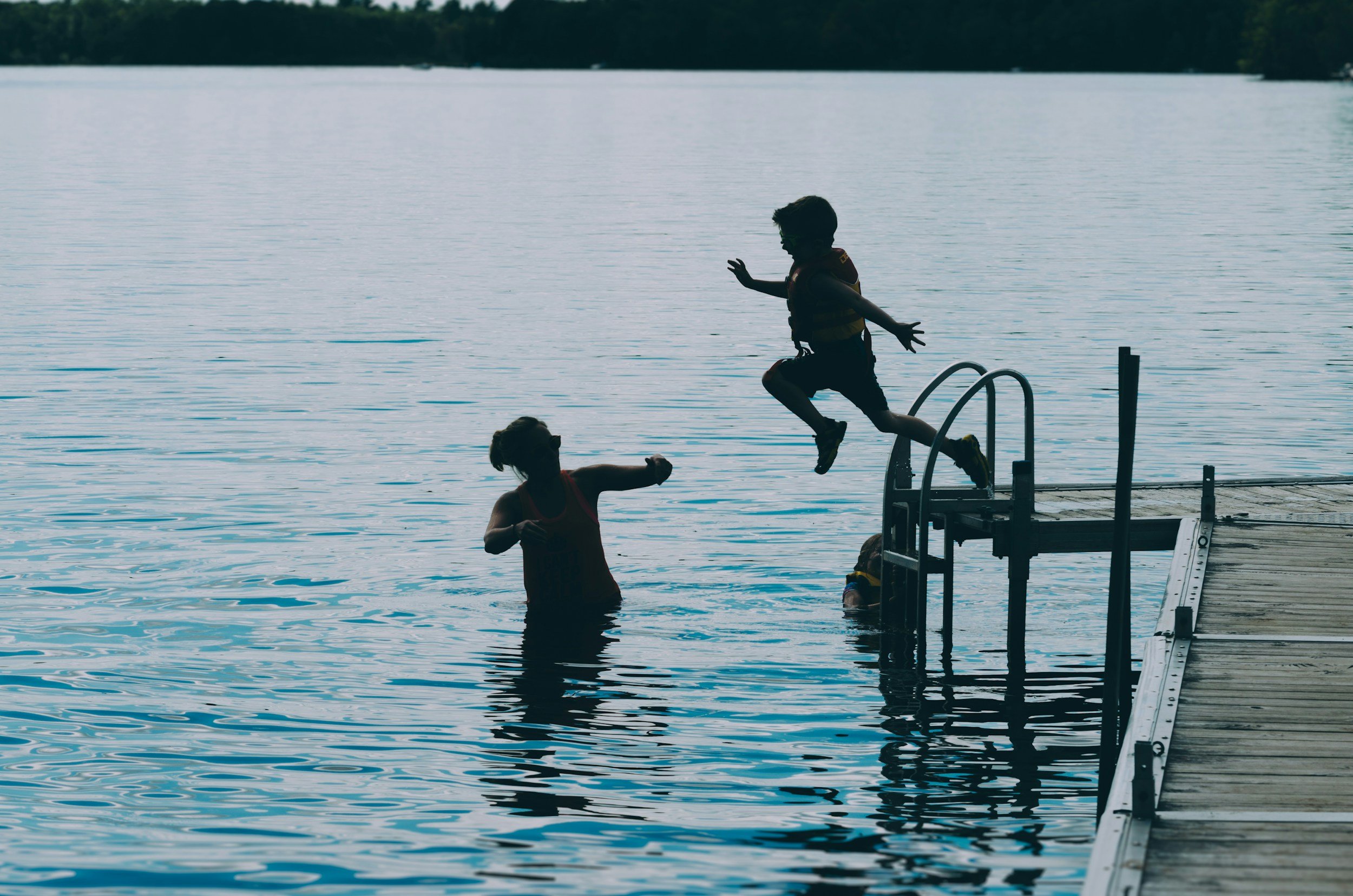 Child jumping off dock into water near adult on a lake.