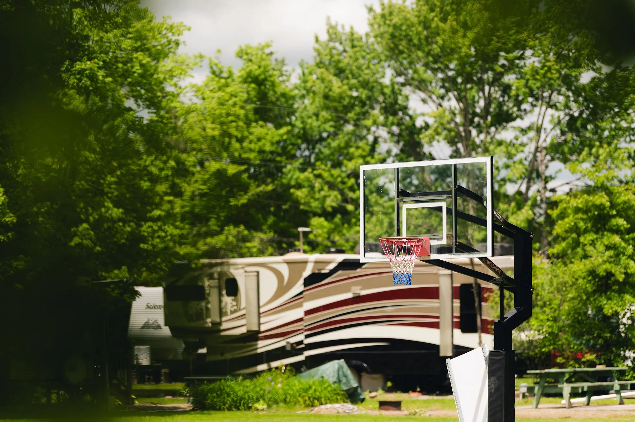 Outdoor basketball hoop on a court with a large RV and green trees in the background.