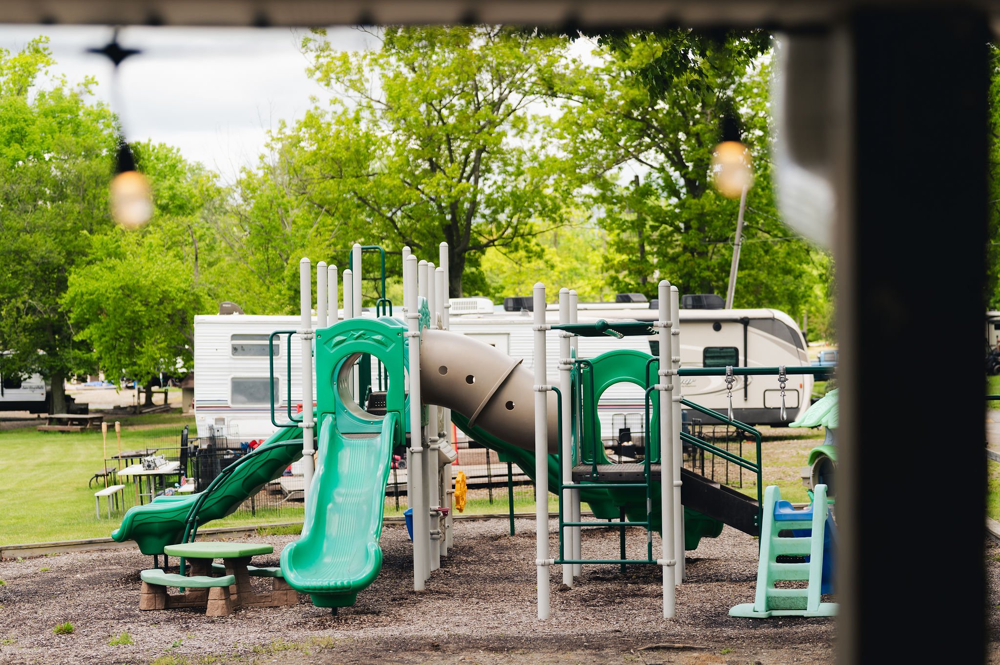 Children's playground with green slides and climbing structures, surrounded by trees and RVs.