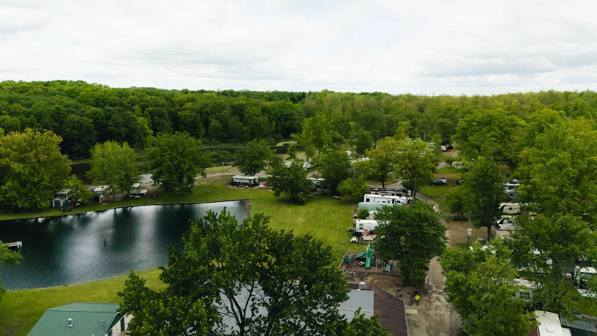 Aerial view of a campground with RVs, trees, and a pond, surrounded by lush green forest and cloudy sky.