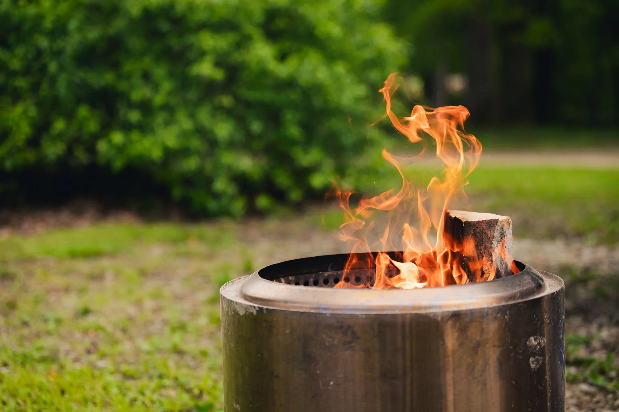 A metal outdoor fire pit with flames burning inside, set in a grassy area with trees in the background.