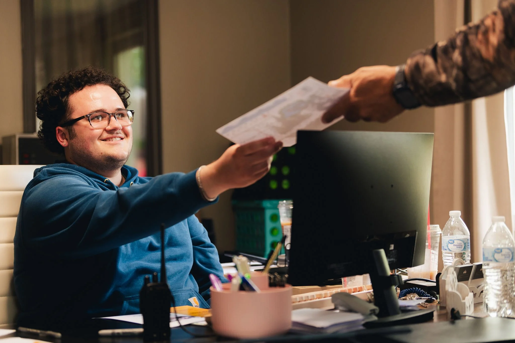 A young man in glasses and a blue hoodie smiling and receiving a document from another person in an office setting.