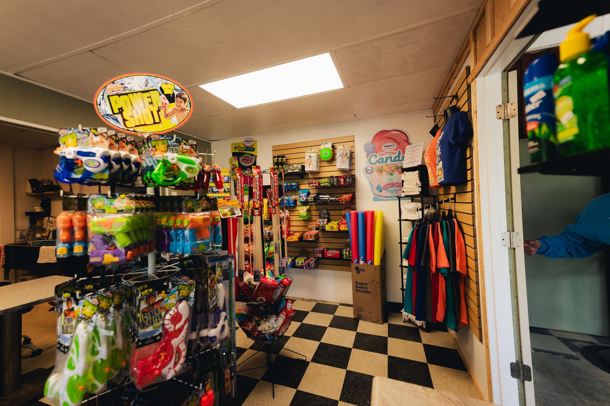 Inside a small store, shelves displaying water guns, pool noodles, and various candy, with clothing items hanging on one wall and a person partially visible at the doorway.