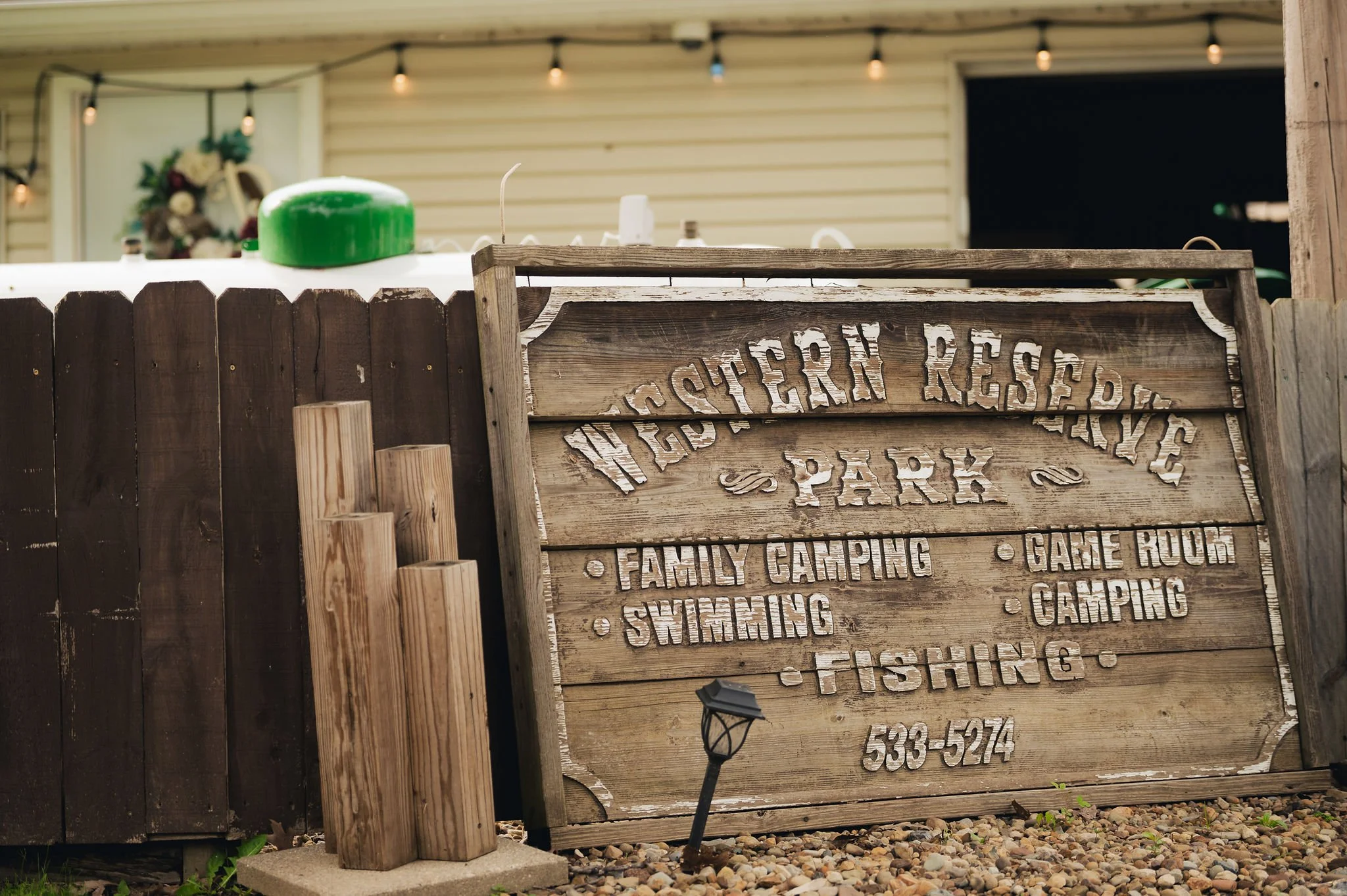 A wooden sign for Western Reserve Park listing activities including family camping, swimming, fishing, game room, and camping with a phone number. There are small wooden posts and pebbles in front of the sign, and a small solar lamp is placed on the 
