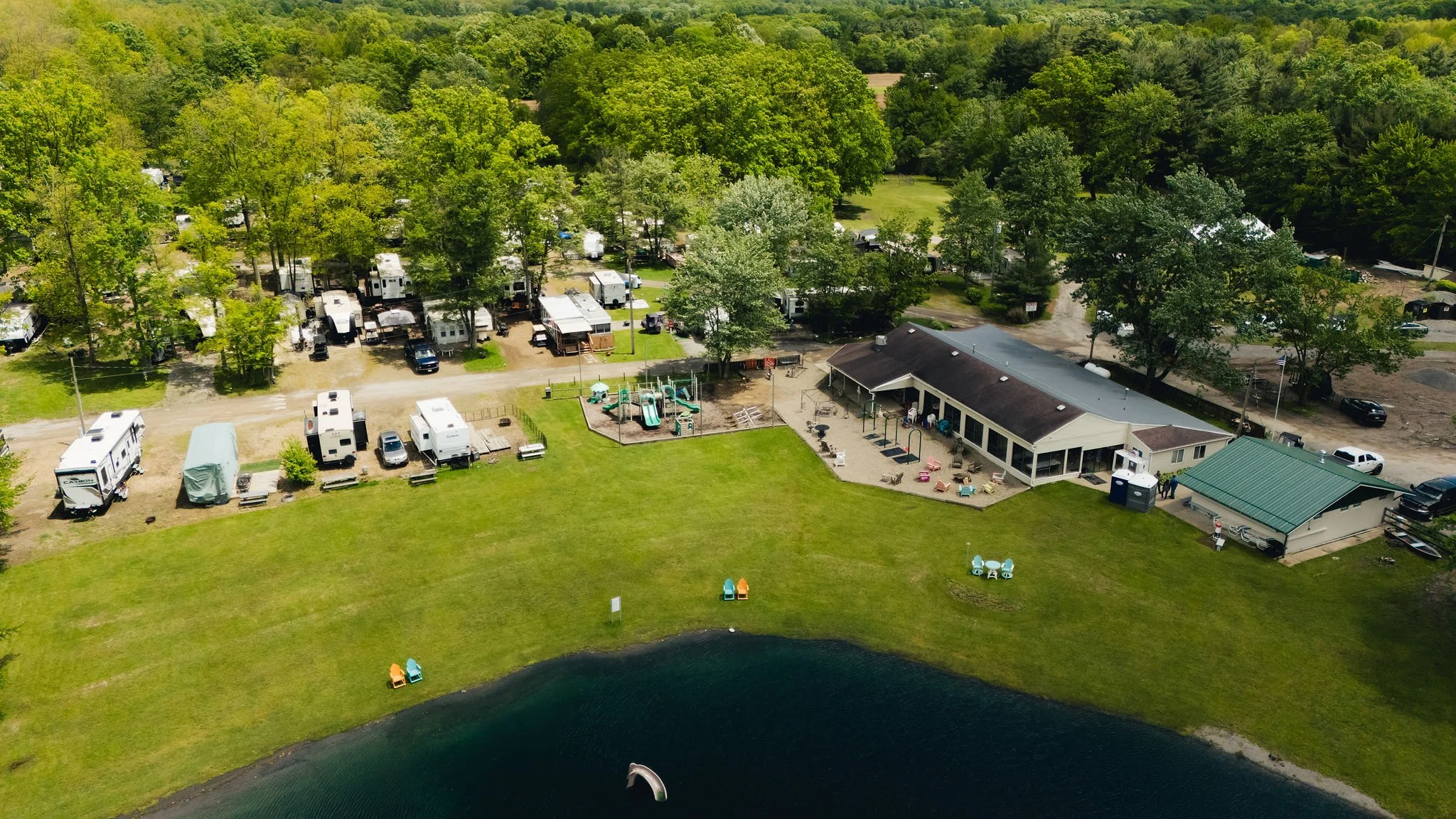 Aerial view of a campground near a lake, with multiple RVs and trailers parked among trees, a playground, a building with outdoor seating, and colorful chairs on the lawn.