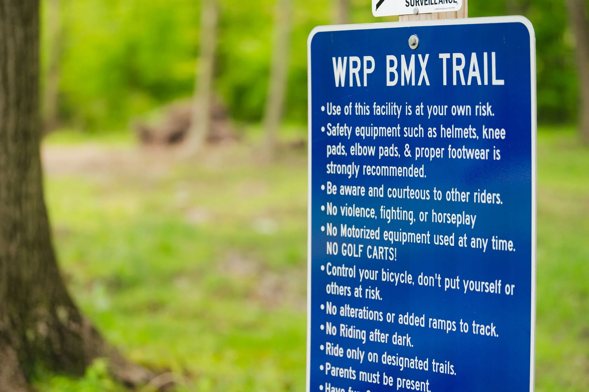 Blue trail sign with safety guidelines for the WRP BMX Trail, including helmet use, courteous riding, no violence or motorized equipment, controlling bicycles, and riding only on designated trails.