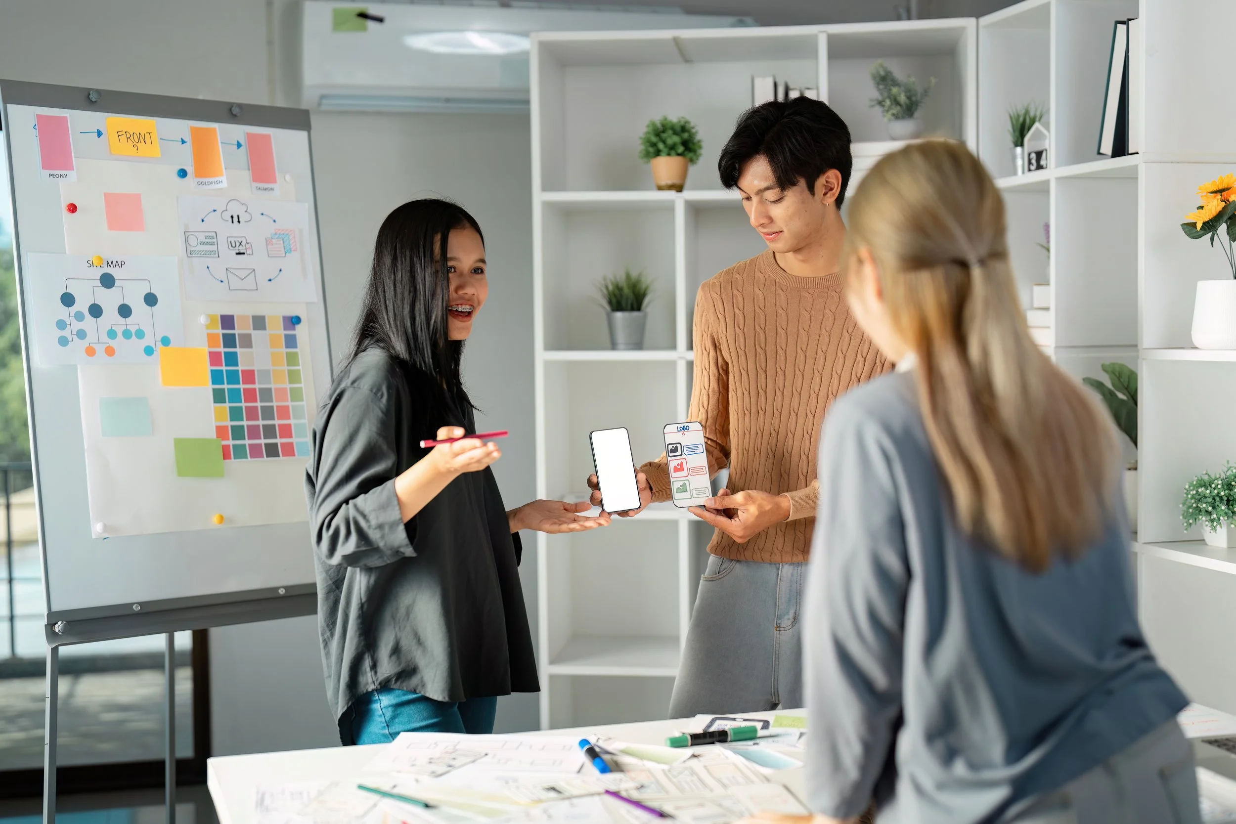 Three people having a discussion in an office setting, with a woman holding a smartphone displaying a blank screen. A man is showing a paper with app design icons. A flip chart with diagrams and color swatches is visible in the background.
