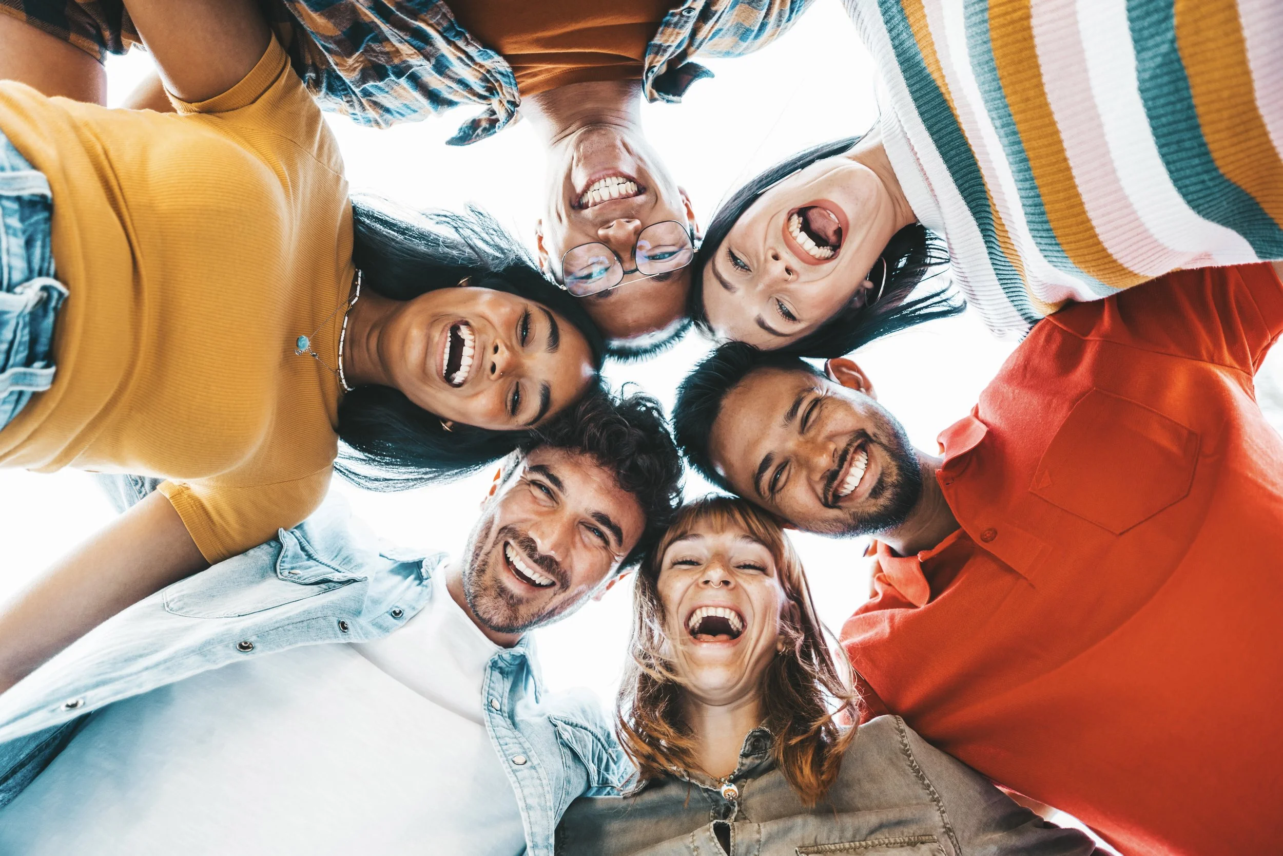 Group of six people standing in a circle looking down at the camera with wide smiles and laughter.