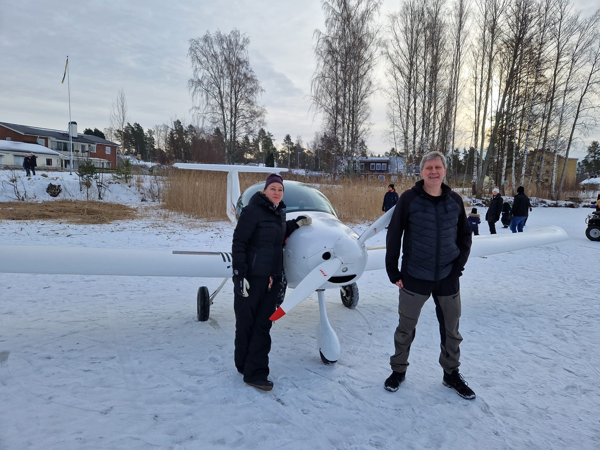 Två personer står framför ett litet flygplan på en snötäckt yta, omgivna av vintriga träd och ett hus i bakgrunden. Flera personer syns längre bort.