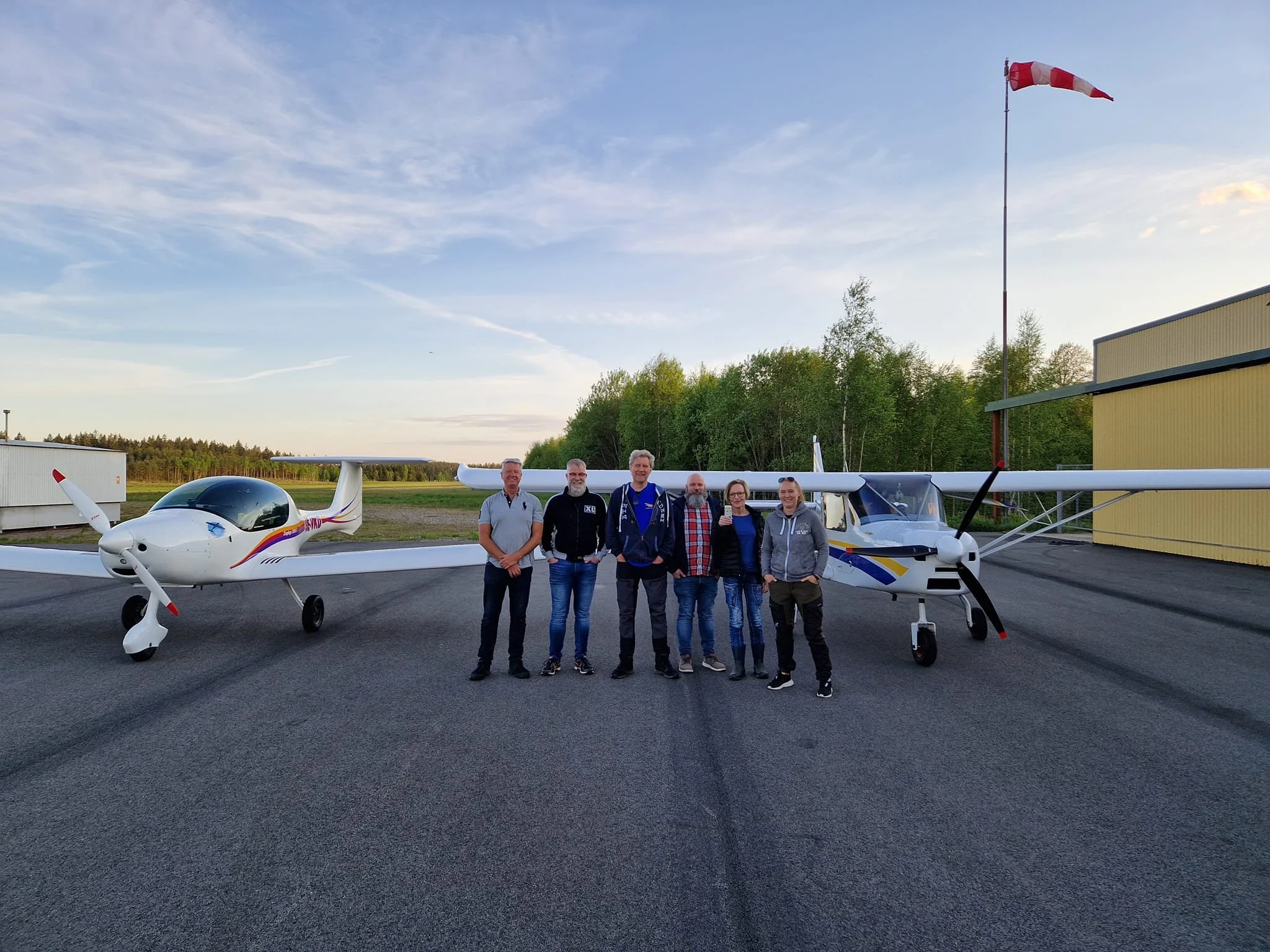 En grupp människor står framför två små flygplan på en flygplats med en blå himmel i bakgrunden.