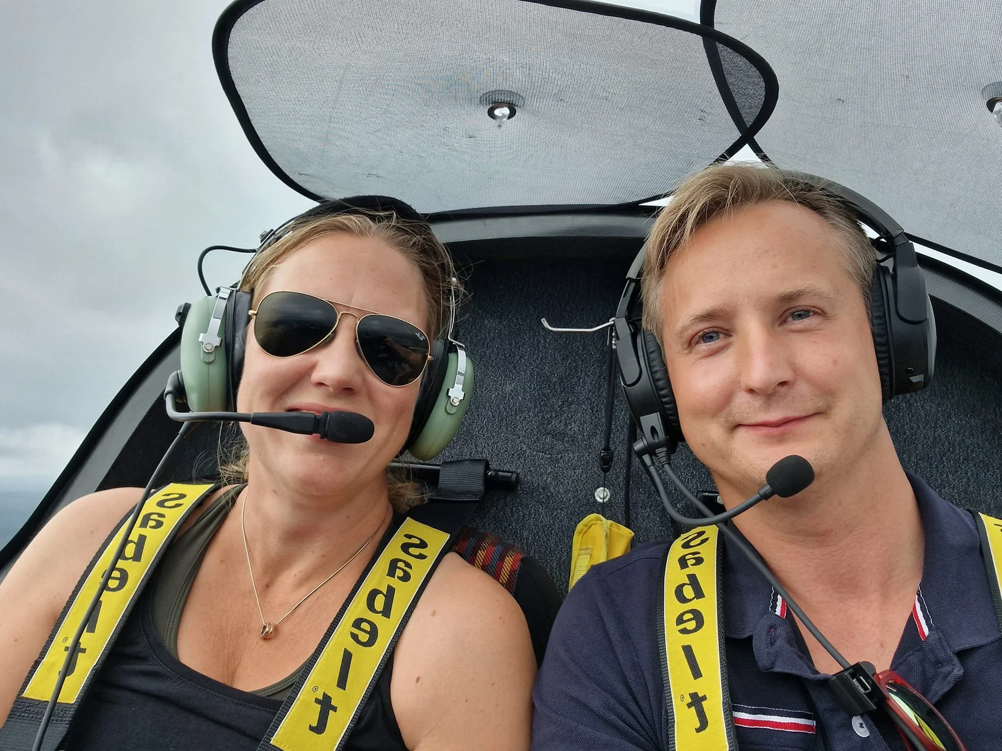 Två personer sitter i cockpit på ett flygplan, med hörlurar och mikrofoner. De bär säkerhetsbälten med texten "selflets." En kvinna till vänster har solglasögon, och mannen till höger ler. Det är molnigt utanför.