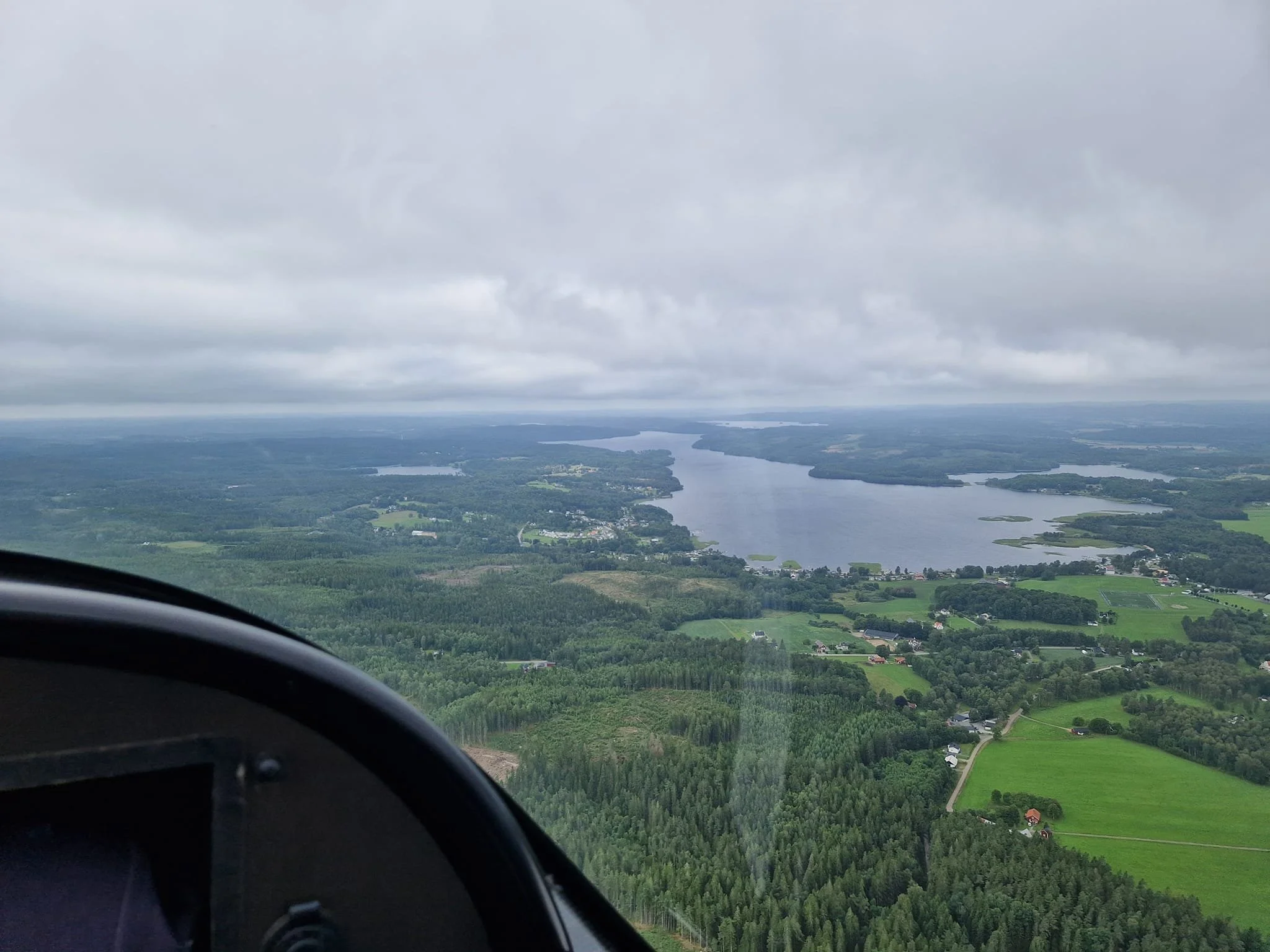 Utsikt från ett flygplan över ett landskap med sjö och skog under en molnig himmel.