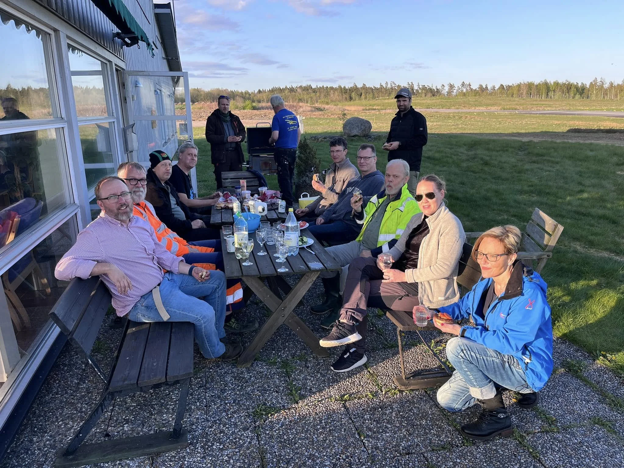 En grupp människor sitter runt ett bord utomhus och har en måltid. Flera personer är klädda i varselkläder och det är en solig dag. Landskapet i bakgrunden visar ett öppet fält.