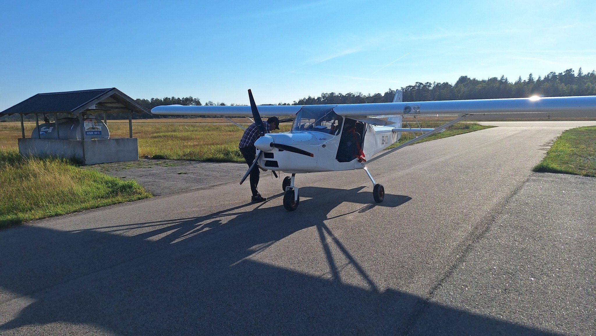 Litet flygplan på landningsbana, person intill flygplanet, klarblå himmel, gräsfält runtom.