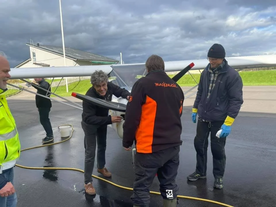 Flera människor arbetar med ett litet flygplan utomhus. De bär varma kläder och verkar engagera sig i underhåll eller rengöring av planet. Himlen är molnig och marken ser blöt ut.