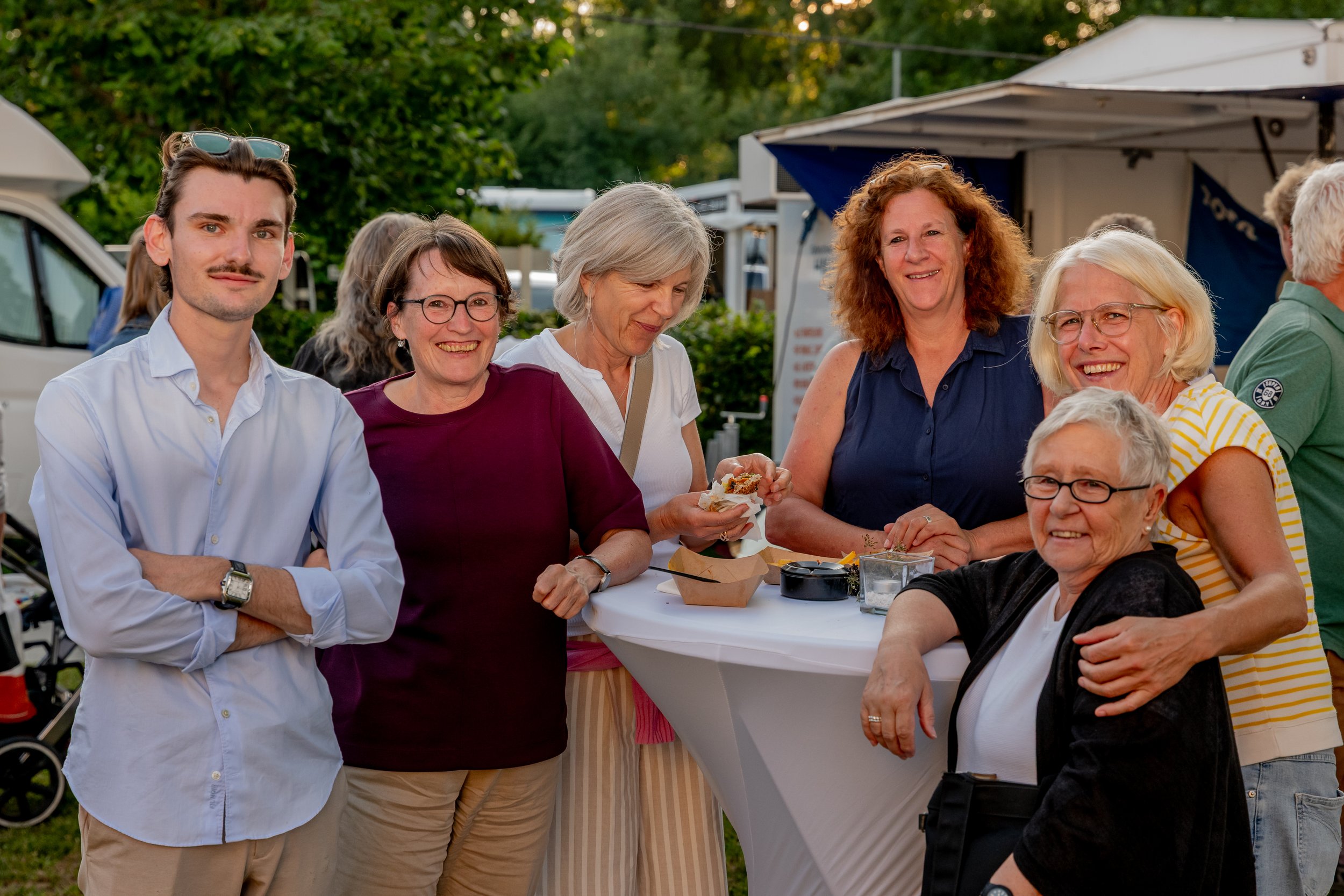 Eine Gruppe von sieben Menschen, bestehend aus älteren Frauen, einem jüngeren Mann und einer älteren Frau, die auf einem Fest im Freien an einem Stehtisch lachen und sich unterhalten.